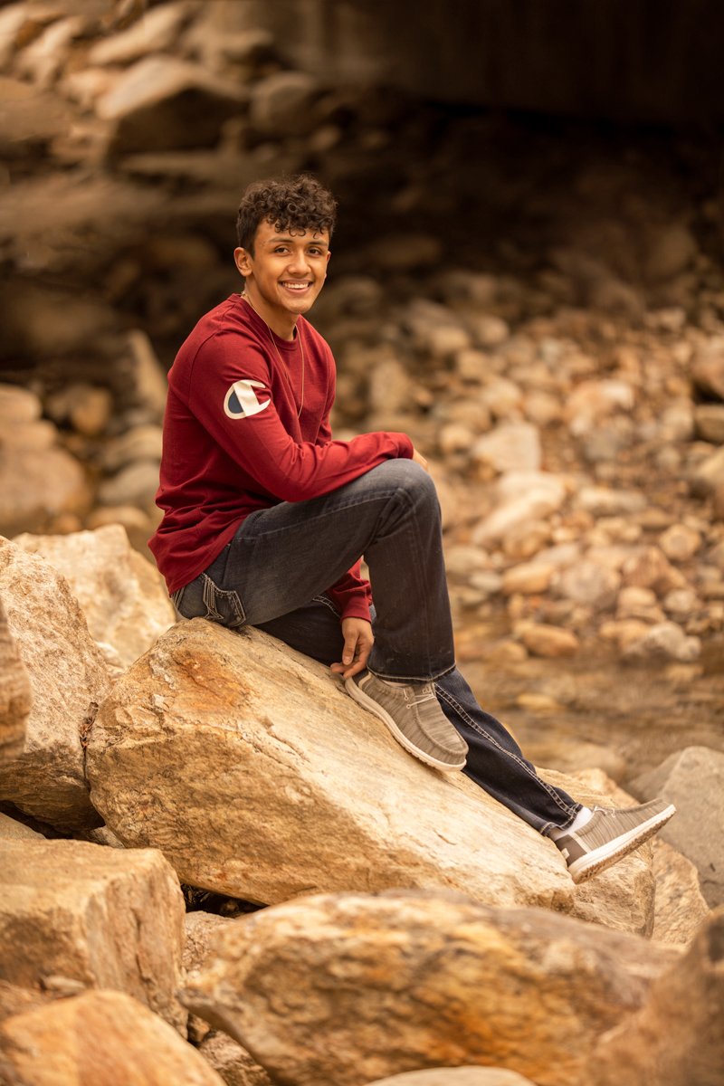 A young man with curly hair sitting on a large rock in a rocky outdoor area, smiling at the camera, wearing a red long-sleeve shirt, black jeans, and beige sneakers.