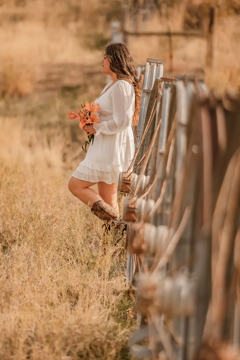 A woman in a white dress holding orange flowers, leaning against a wooden fence in a field with tall grass, during sunset or golden hour.