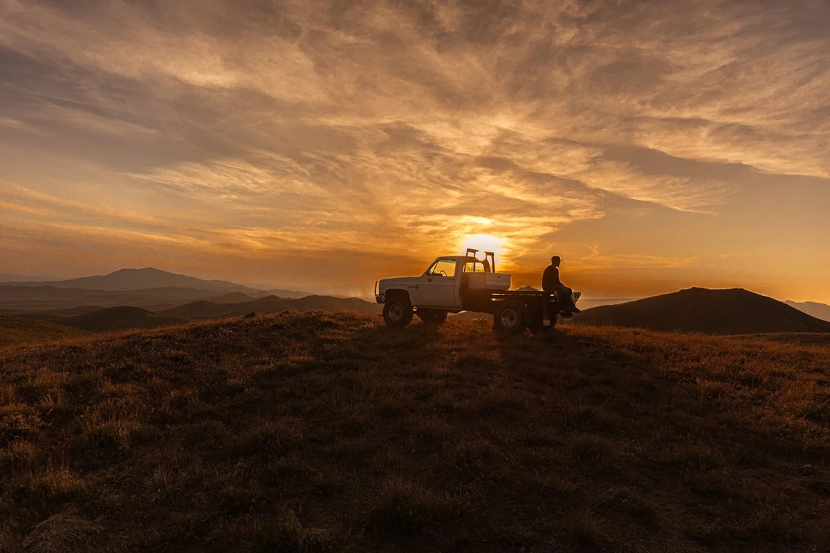 A person sitting on the side of a white pickup truck parked on a grassy hilltop during sunset with a mountain range in the distance and a sky filled with colorful clouds.