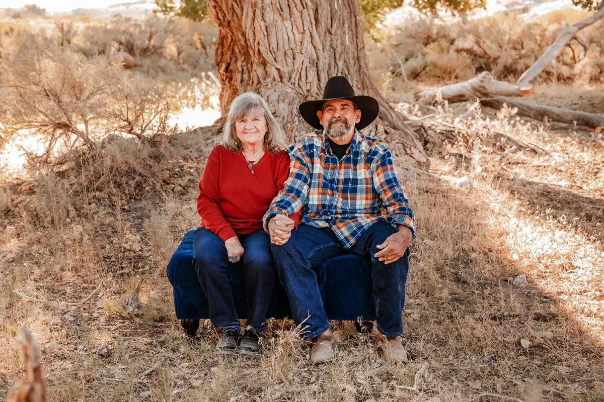 A smiling elderly woman and an elderly man with a gray beard sitting on a dark blue velvet bench outdoors in a natural setting with dry grass, bushes, and a large tree, holding hands. The woman wears a red sweater and jeans, and the man wears a plaid