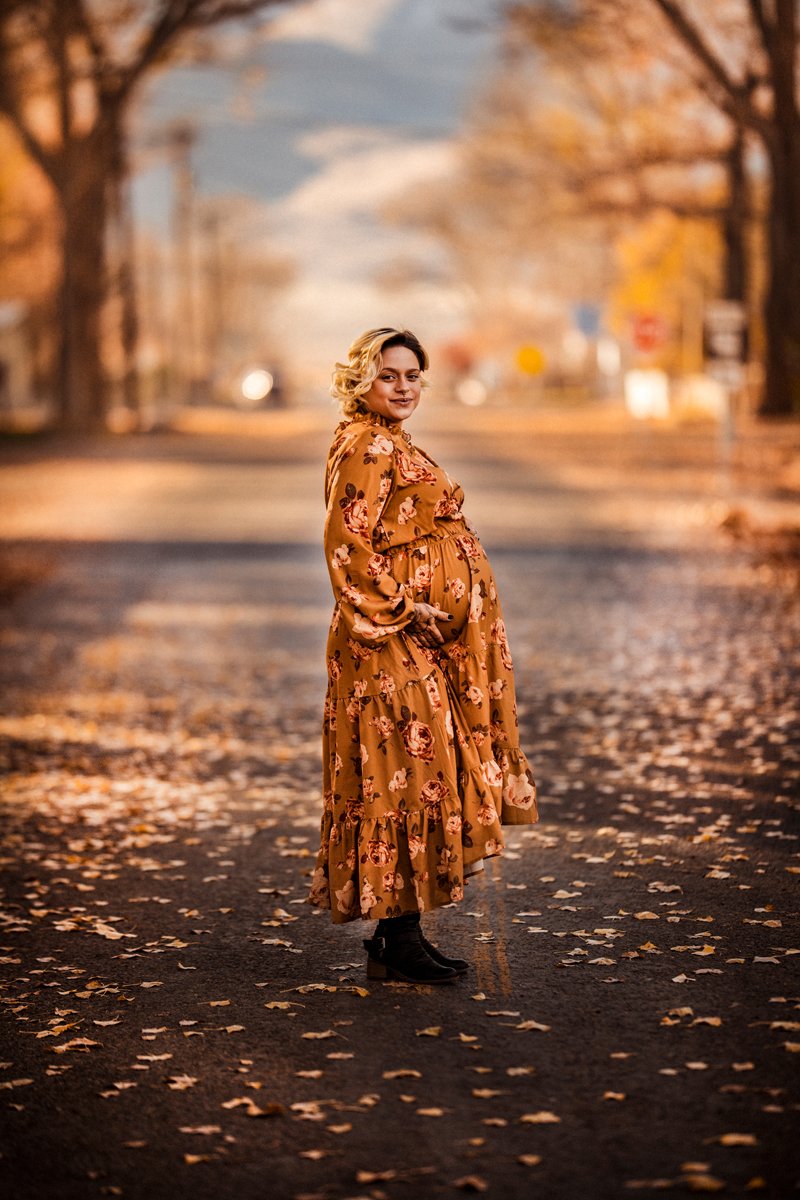 A pregnant woman in a long floral dress standing on a fall-themed street with autumn leaves on the ground and trees with orange leaves in the background.