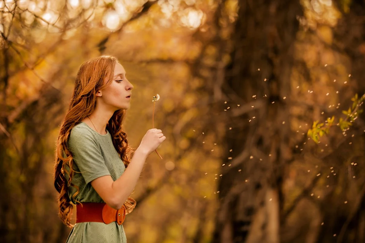 A woman with long red hair in a green dress holding a dandelion in a forest with autumn foliage.