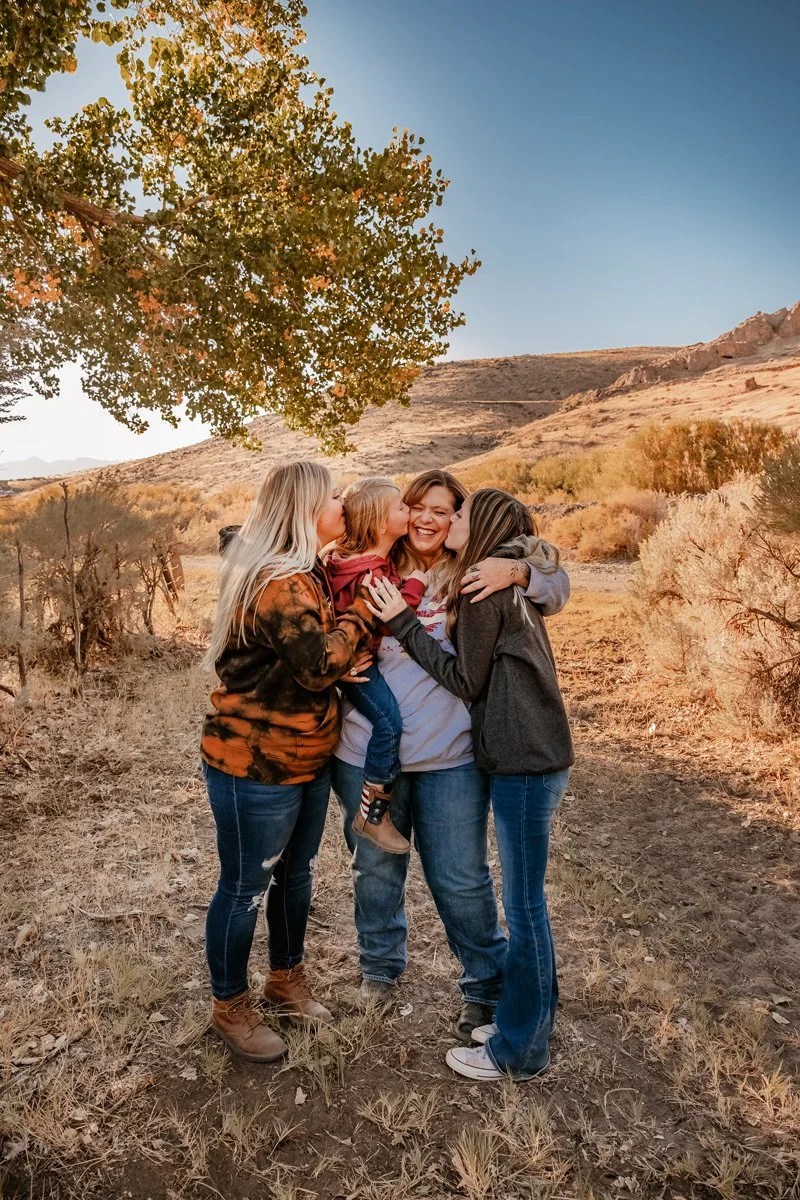 Four women and a young girl sharing a group hug and kisses outdoors in a desert landscape with a large green tree and hills in the background during sunset.