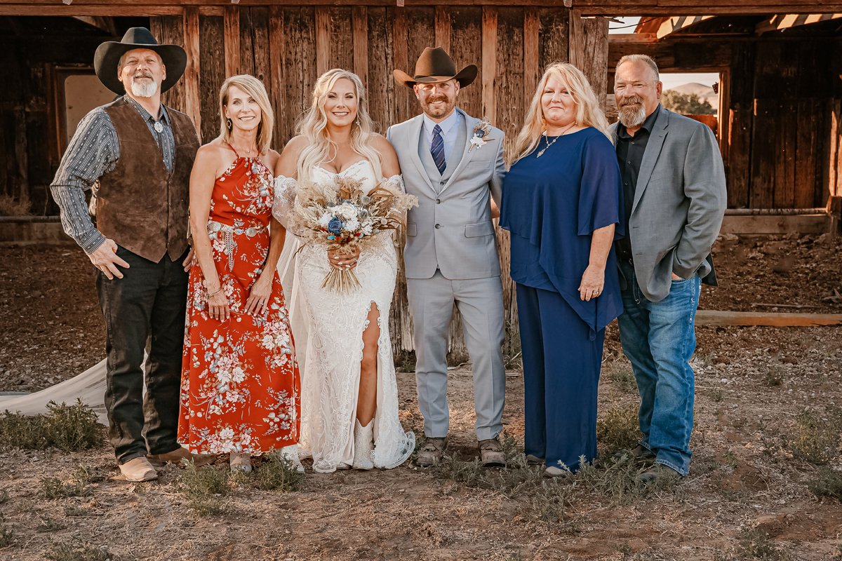 Group of six people standing outdoors in front of a wooden barn, dressed in wedding and casual attire, celebrating a wedding with a bride holding a bouquet and everyone smiling.