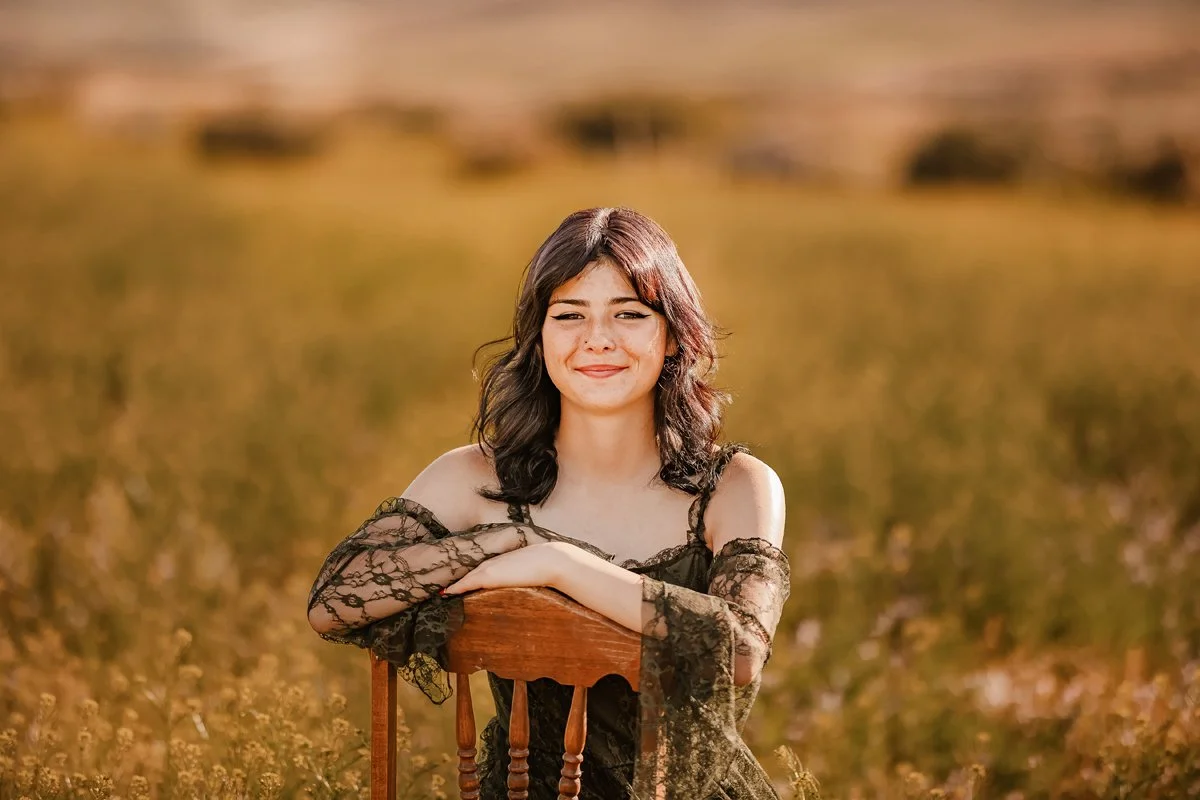 A young woman sitting on a wooden chair outdoors in a field with tall grasses, wearing a black lace dress, smiling gently with one eye closed.
