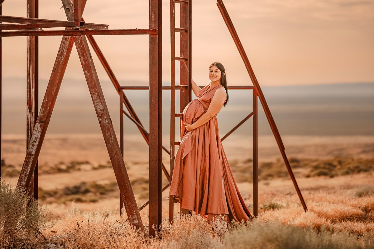 A pregnant woman in a long, flowing dress stands next to an old, rusted metal structure in a desert landscape during sunset, smiling and cradling her belly.