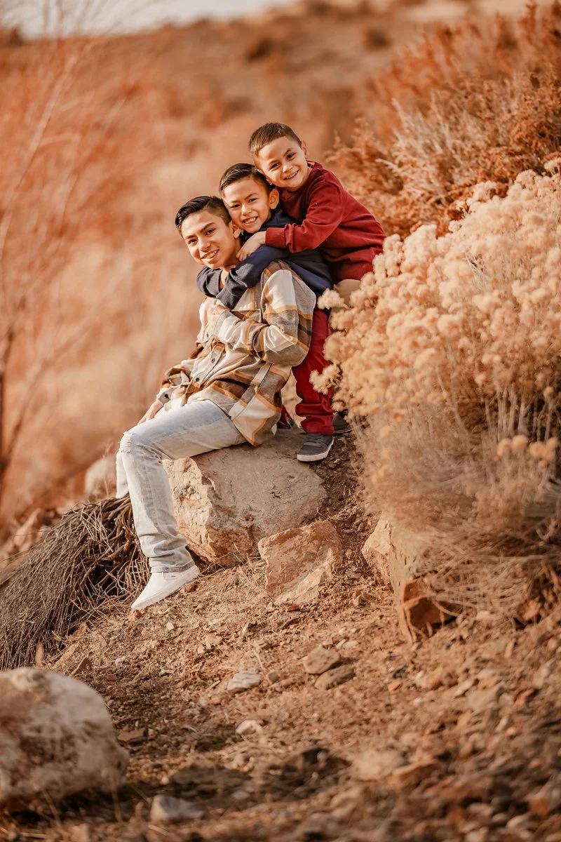 Three boys sitting on a rock outdoors among orange-brown foliage, smiling and embracing each other.