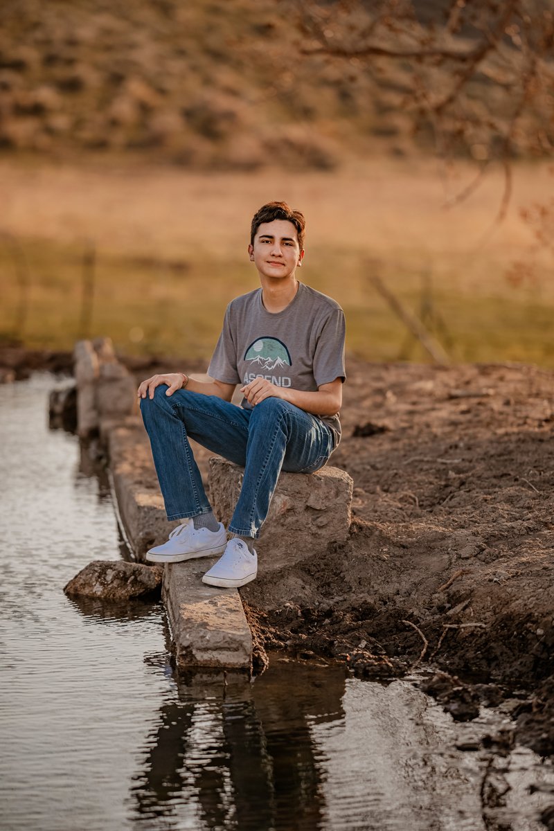 A young man seated on a stone ledge by a body of water during sunset, wearing a gray t-shirt, blue jeans, and white sneakers.