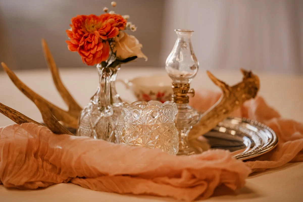Decorative arrangement with a glass vase with red flowers, a small glass oil lamp, a cut glass container, a metal tray, antlers, and pink fabric, all on a table.