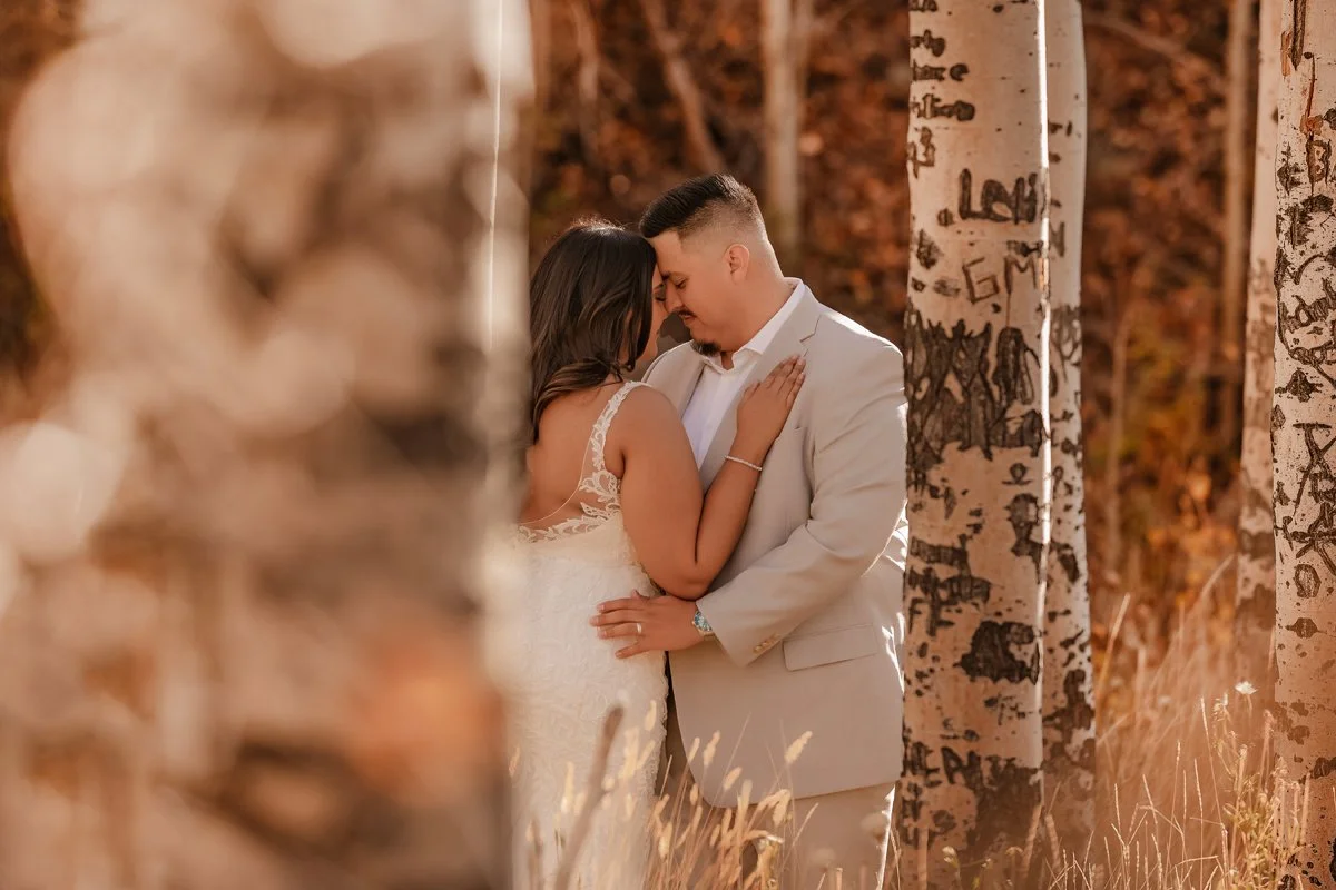 A couple sharing a tender moment in an autumn forest, with their foreheads touching and eyes closed, surrounded by tall, graffiti-covered trees.