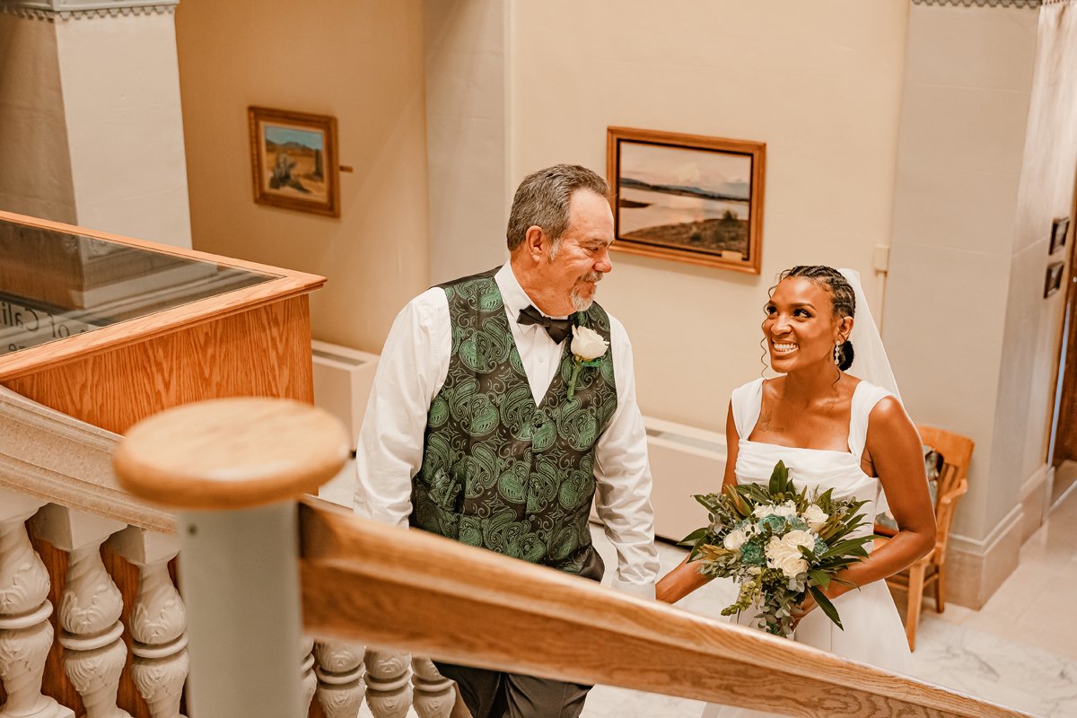 A bride with a bouquet of flowers smiling at an older man with gray hair and a beard, wearing a green vest and black bow tie, on a staircase in an indoor setting.