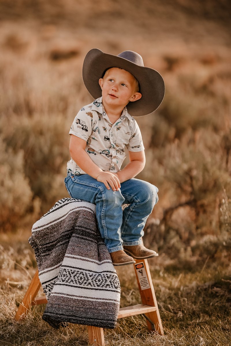 Young boy wearing a cowboy hat and a patterned shirt, sitting on a small wooden stool with a blanket, outdoors in a grassy, desert-like area.