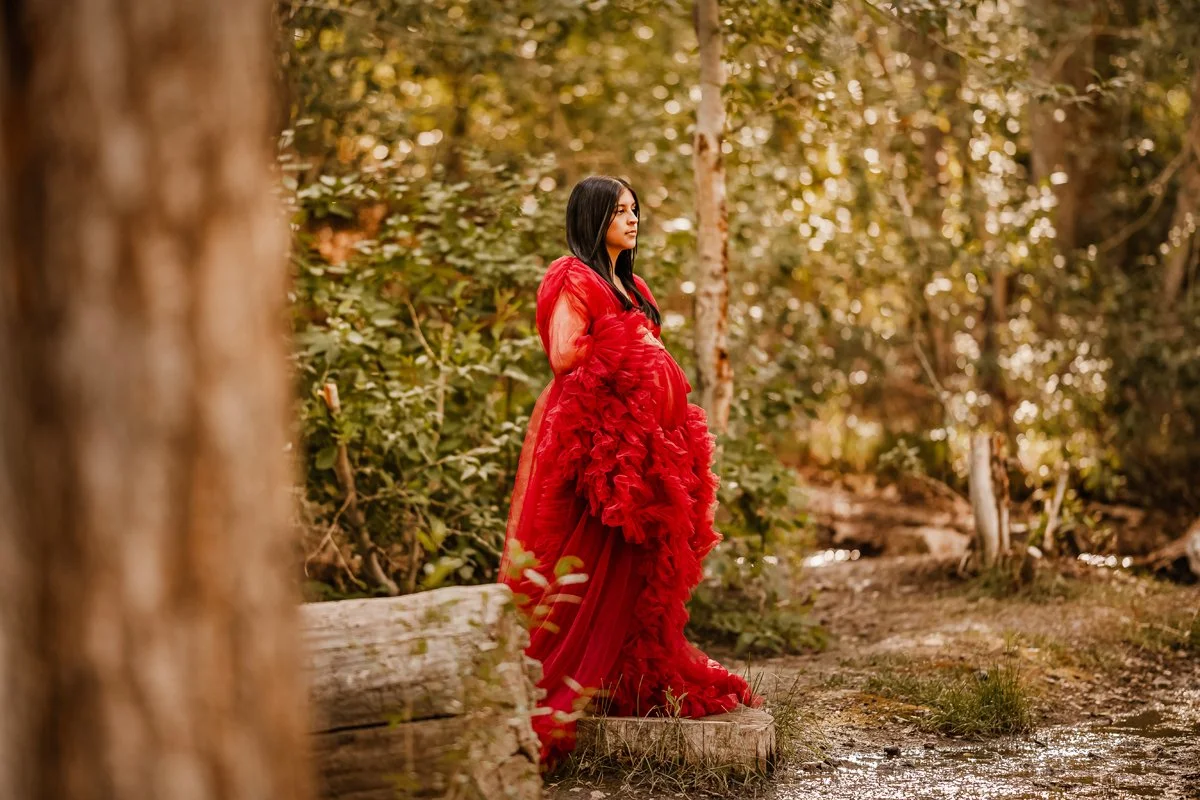 A pregnant woman in a flowing red dress stands on a tree stump in a wooded area during daytime.
