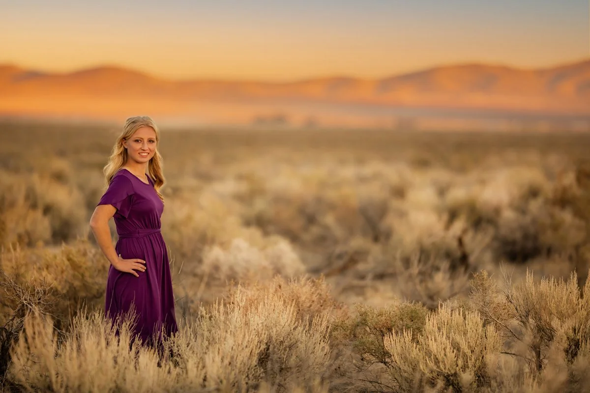 A woman in a purple dress standing in a desert landscape with bushes and mountains in the background during sunset.