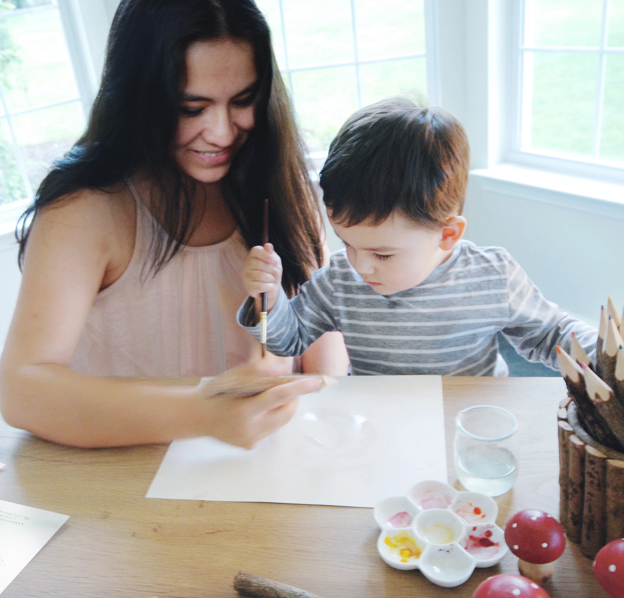 A woman and a young boy sitting at a table, engaging in an outdoor painting activity. The woman is holding a paper, and the boy is using a paintbrush. The table has a palette with paint and some decorative mushrooms, with a window in the background letting in natural light.
