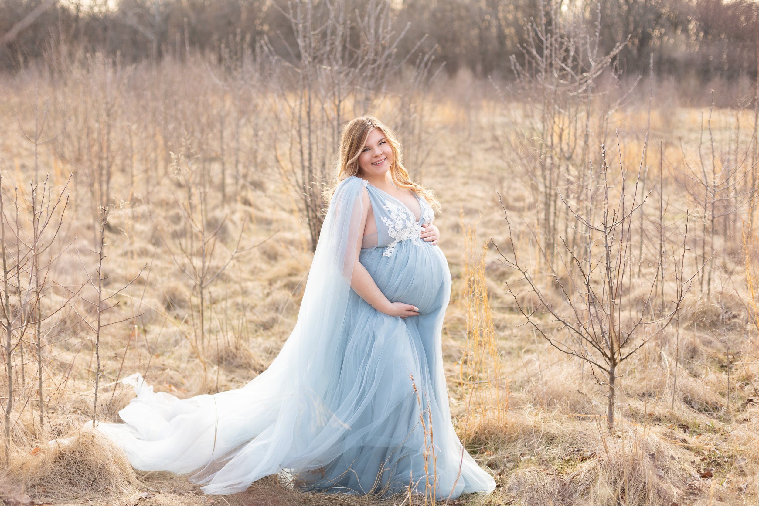 Elegant outdoor maternity portrait featuring an expectant mother in a flowing light blue gown in a field during sunset.