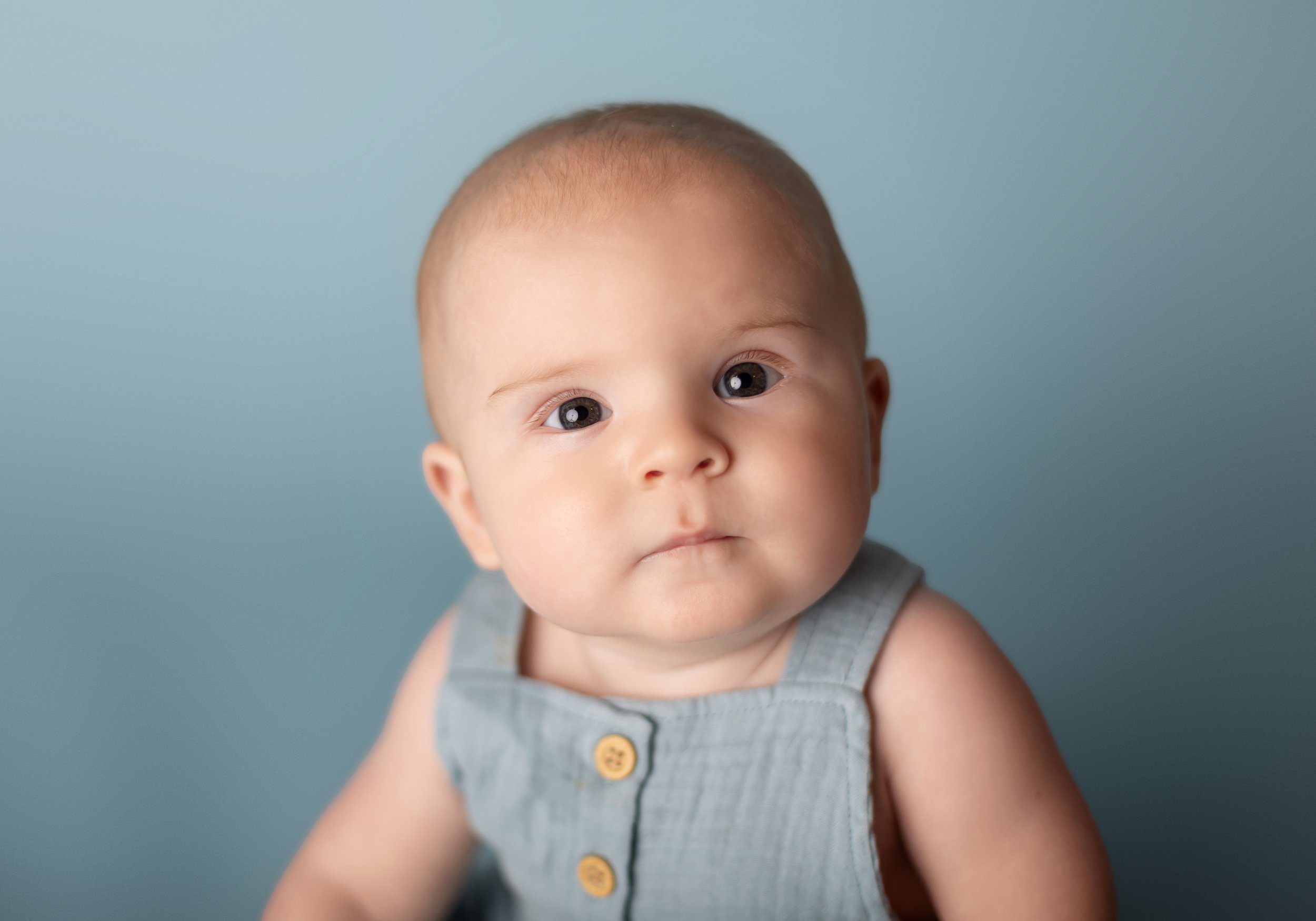 Close up portrait of a baby in a sleeveless denim romper against a soft blue background