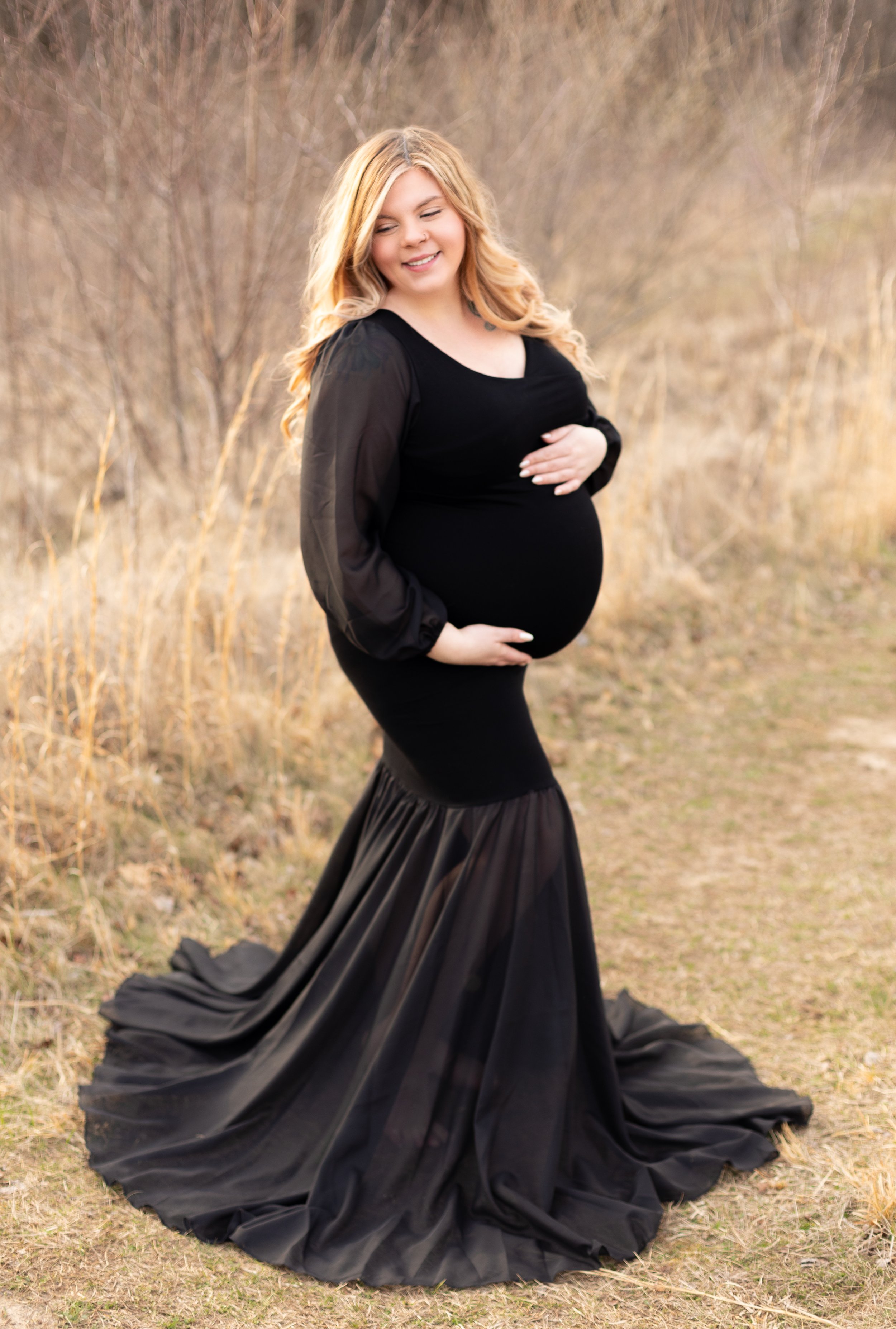 Striking maternity photo of glowing mother to be in a fitted black gown, captured outdoors in a field with a golden hour backdrop.