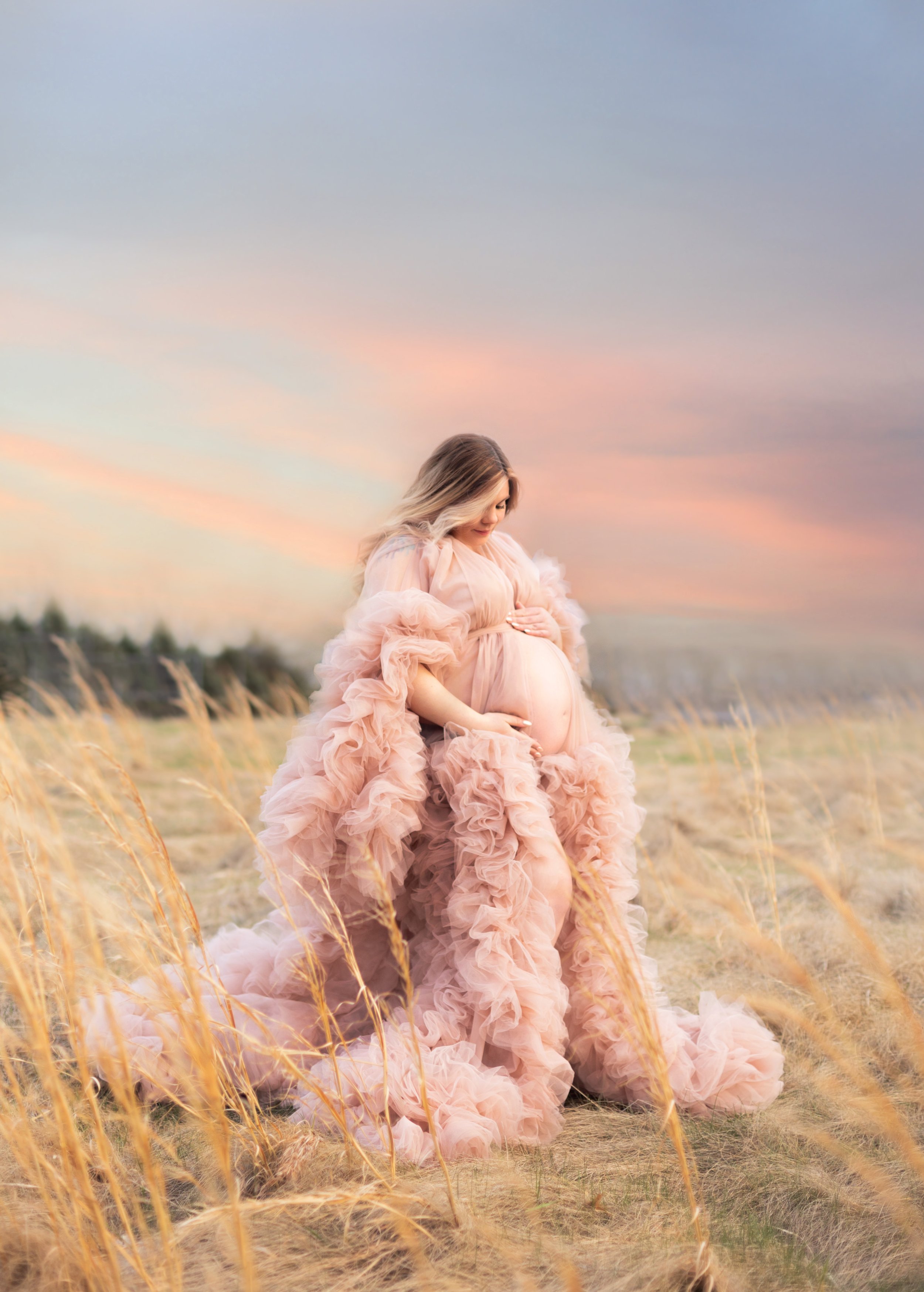 Golden hour maternity photo in a field with expecting mother in a pink ruffle gown with sunset backdrop