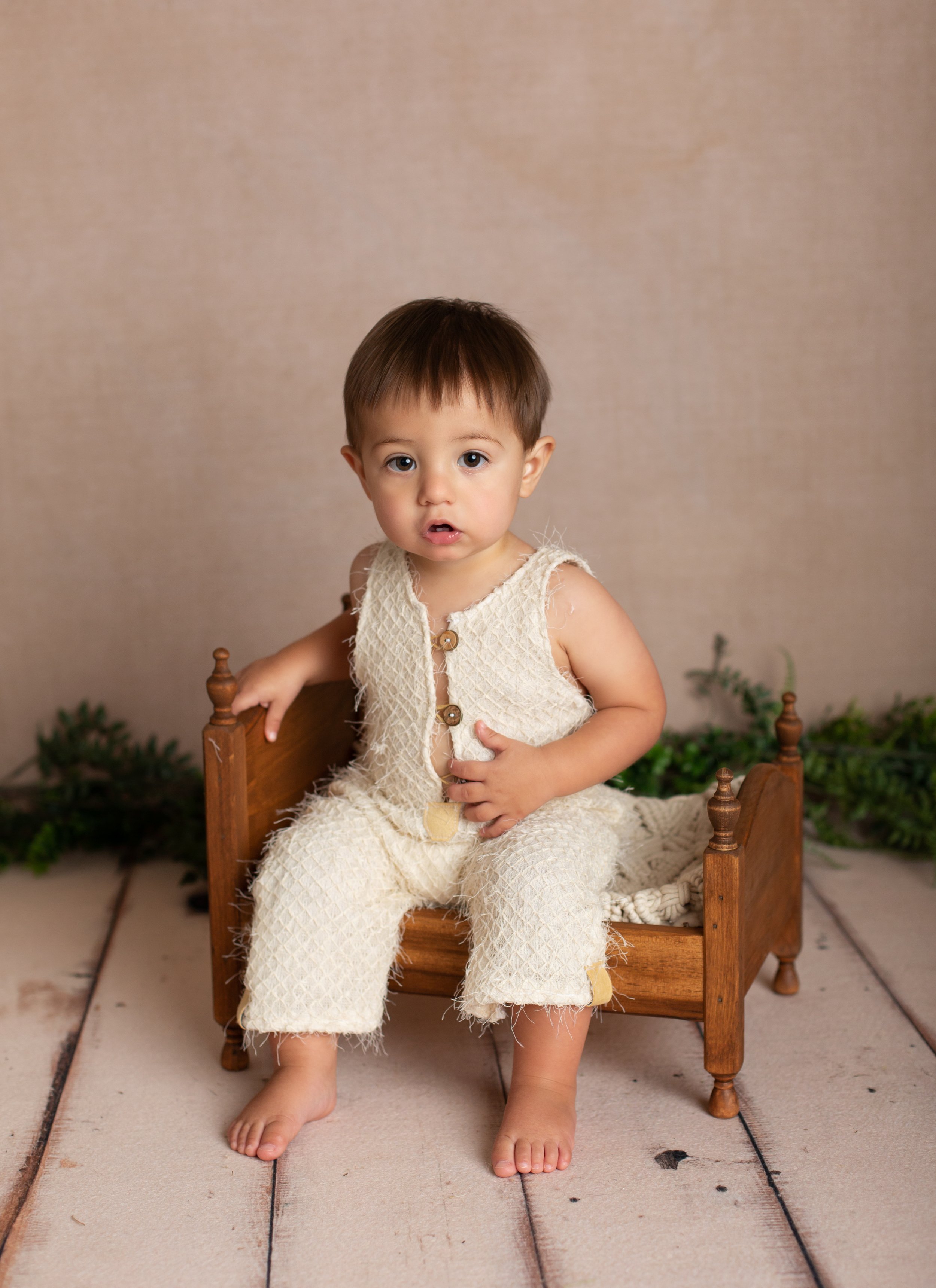 Rustic milestone photo of a toddler sitting on a vintage wooden bench, styled with neutral tones and greenery.