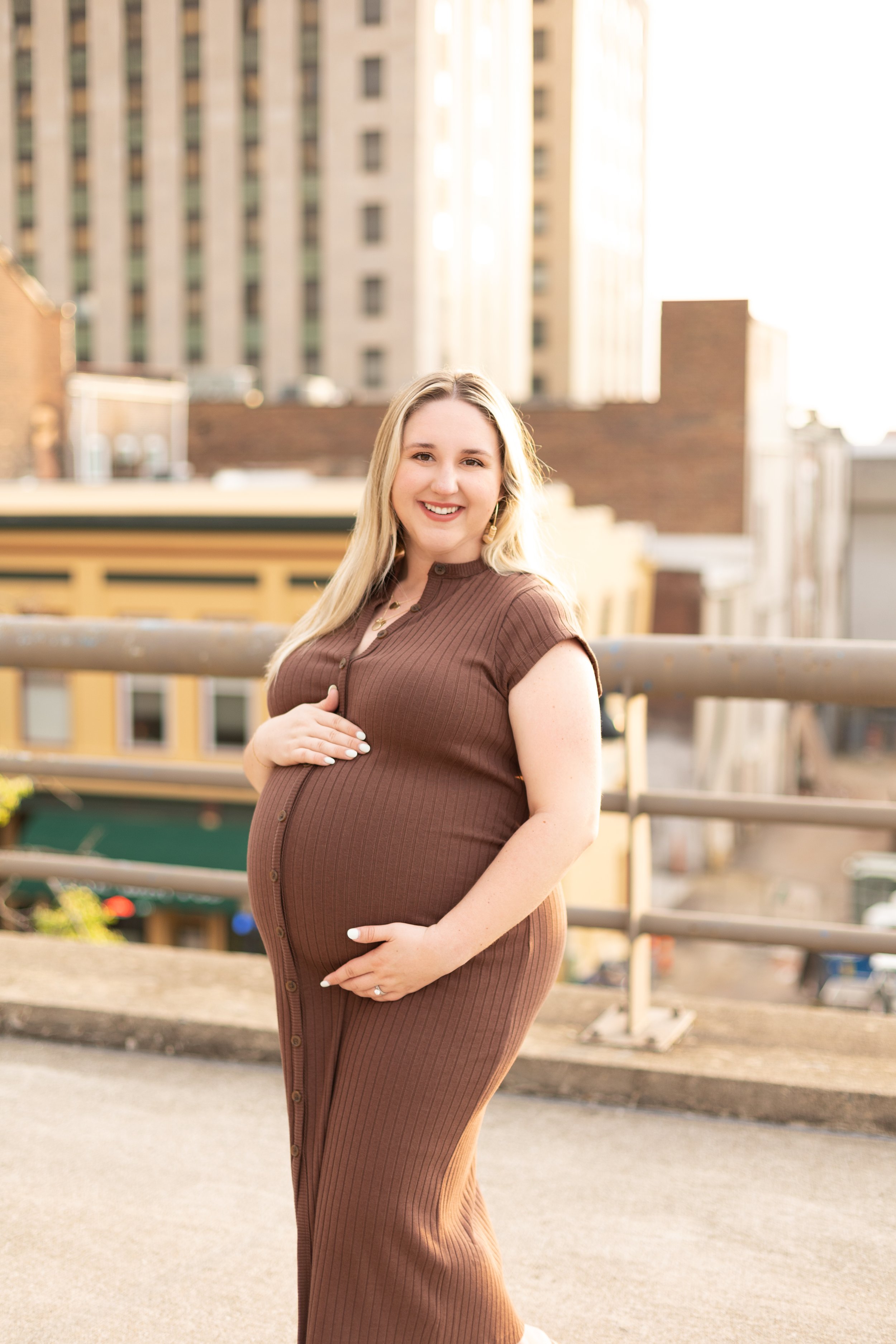 Confident expecting mom posing in a brown fitted dress on a downtown rooftop with city buildings glowing in the background.