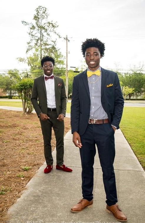  Laron in a classic dark suit and bright-yellow bowtie, and Laroy in a slate-gray suit with a paisley bowtie, smiling and posing in front of a rustic background 