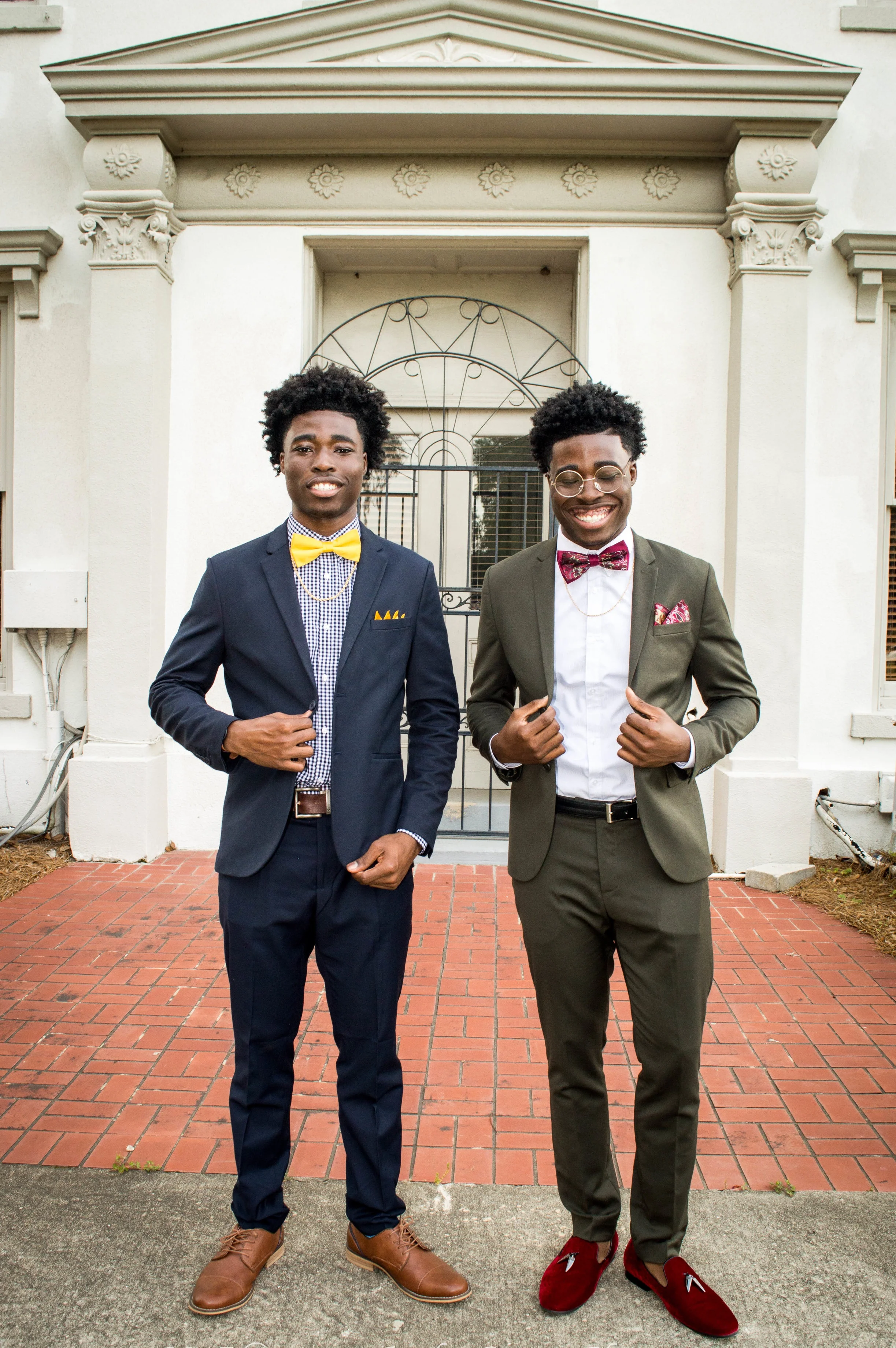  Laron in a classic dark suit and bright-yellow bowtie, and Laroy in a slate-gray suit with a paisley bowtie, smiling and posing in front of a rustic background 