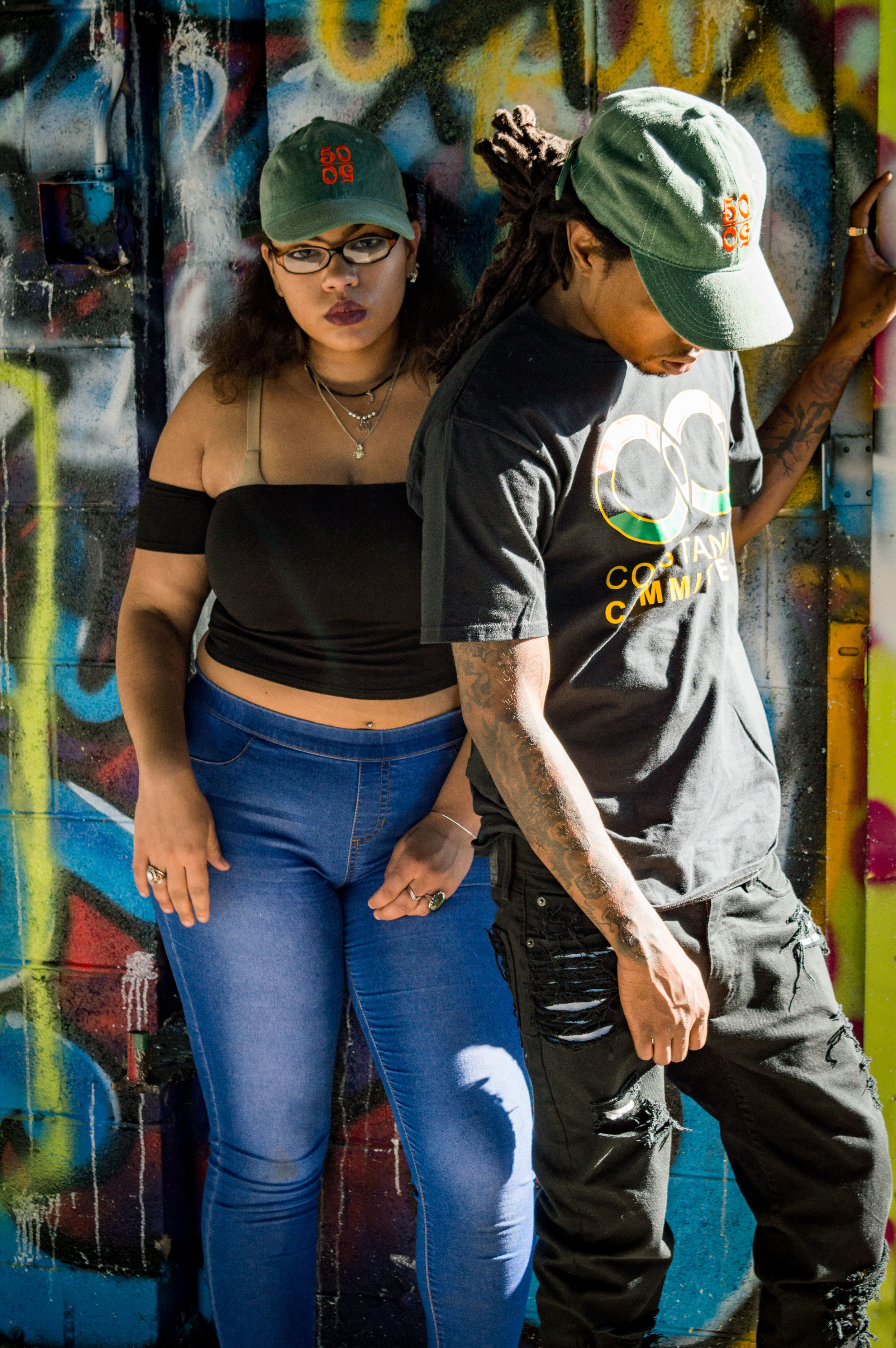  Female model wearing a green trucker hat with a 50/50 logo, posing next to a male model in a another green 50/50 hat, a black shirt, and jeans, in front of a graffiti-covered wall 