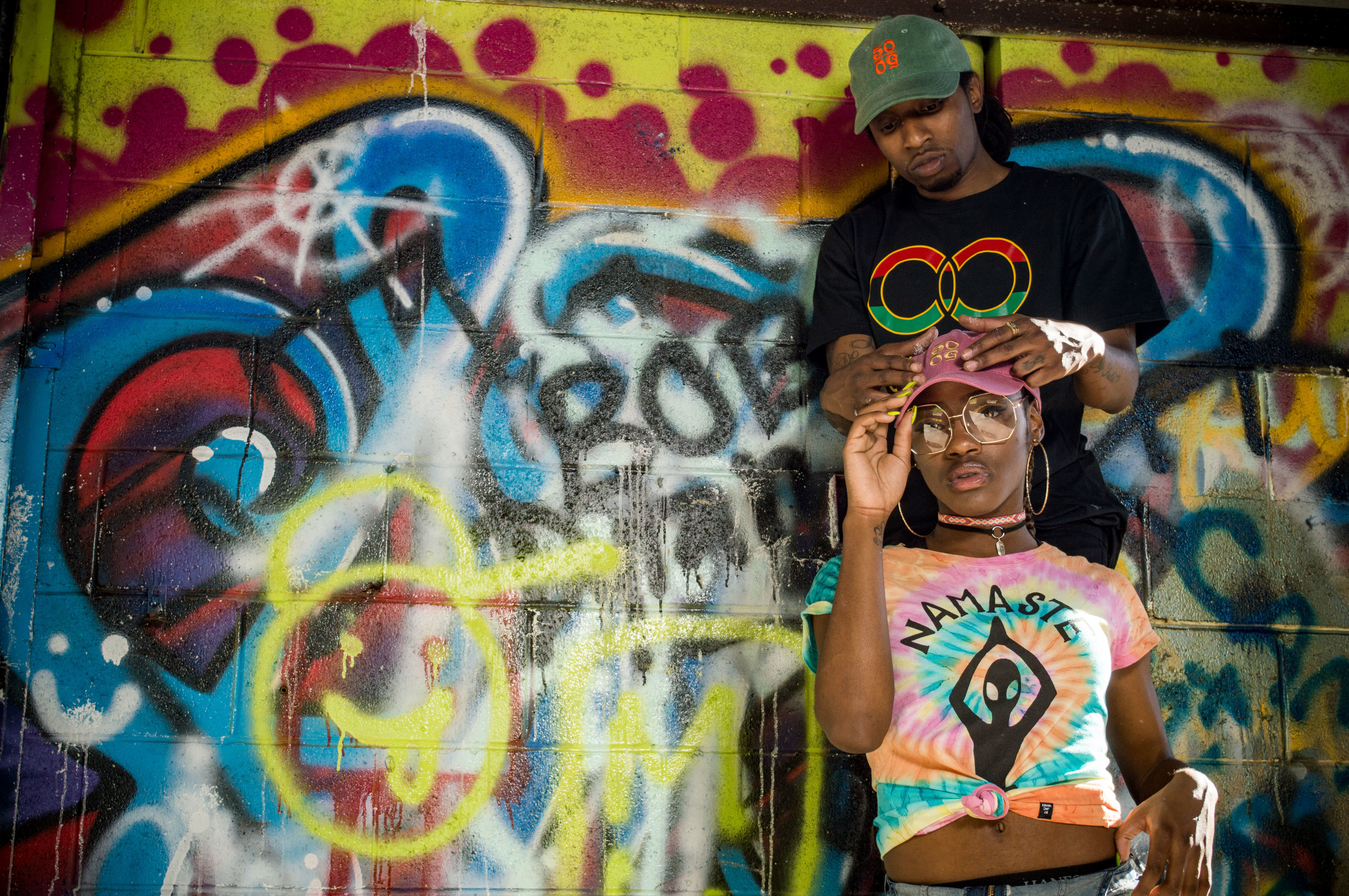  Female model wearing a brightly-colored shirt that says 'namaste' and a pink trucker hat with a 50/50 logo, posing with a male model in a black shirt and green 50/50 hat behind her against a graffiti-covered wall 