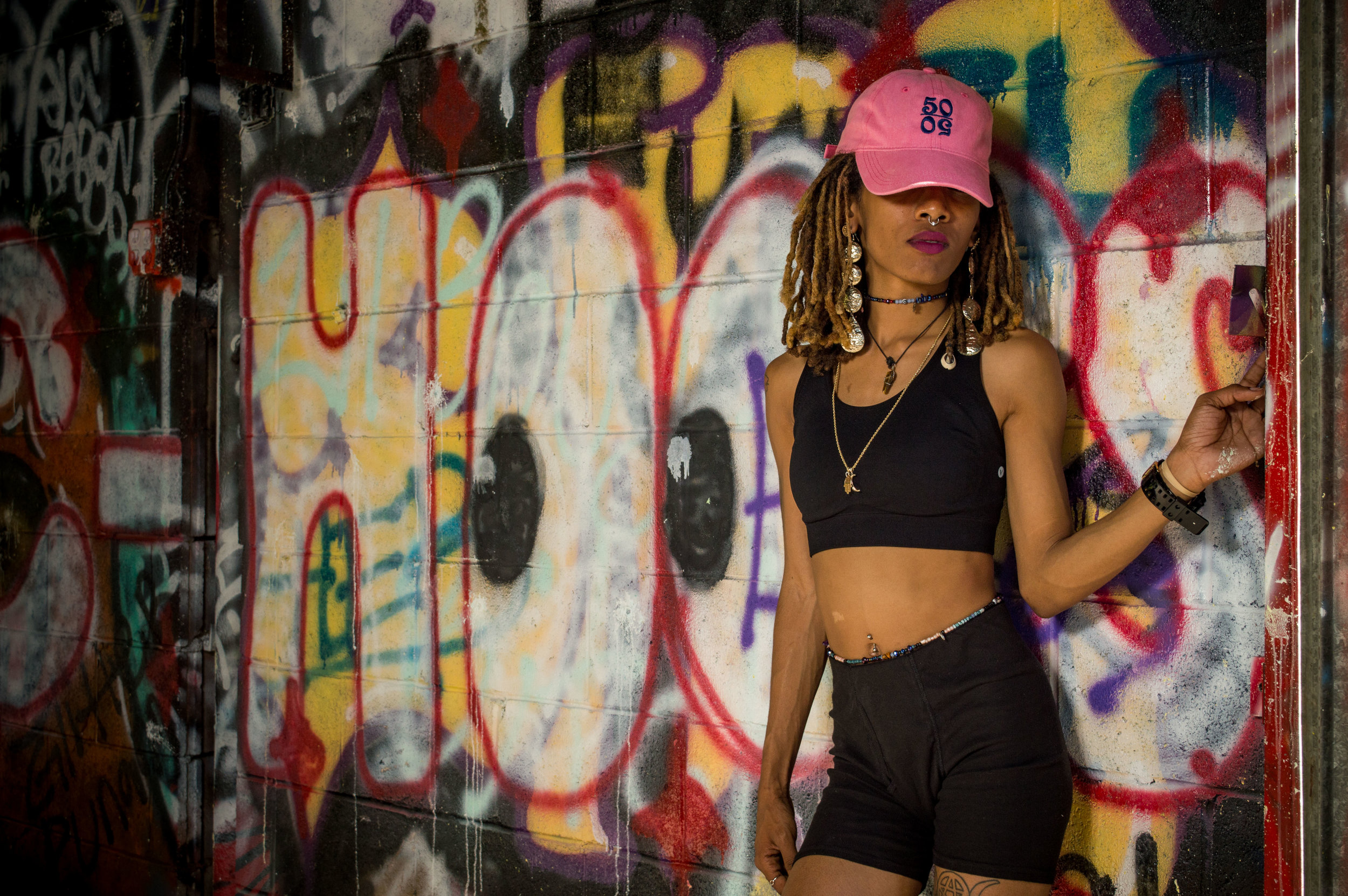  Female model wearing a pink trucker hat with 50/50 logo pulled down low over her eyes, standing in front of a graffiti wall, in a black crop top and high- waisted shorts 