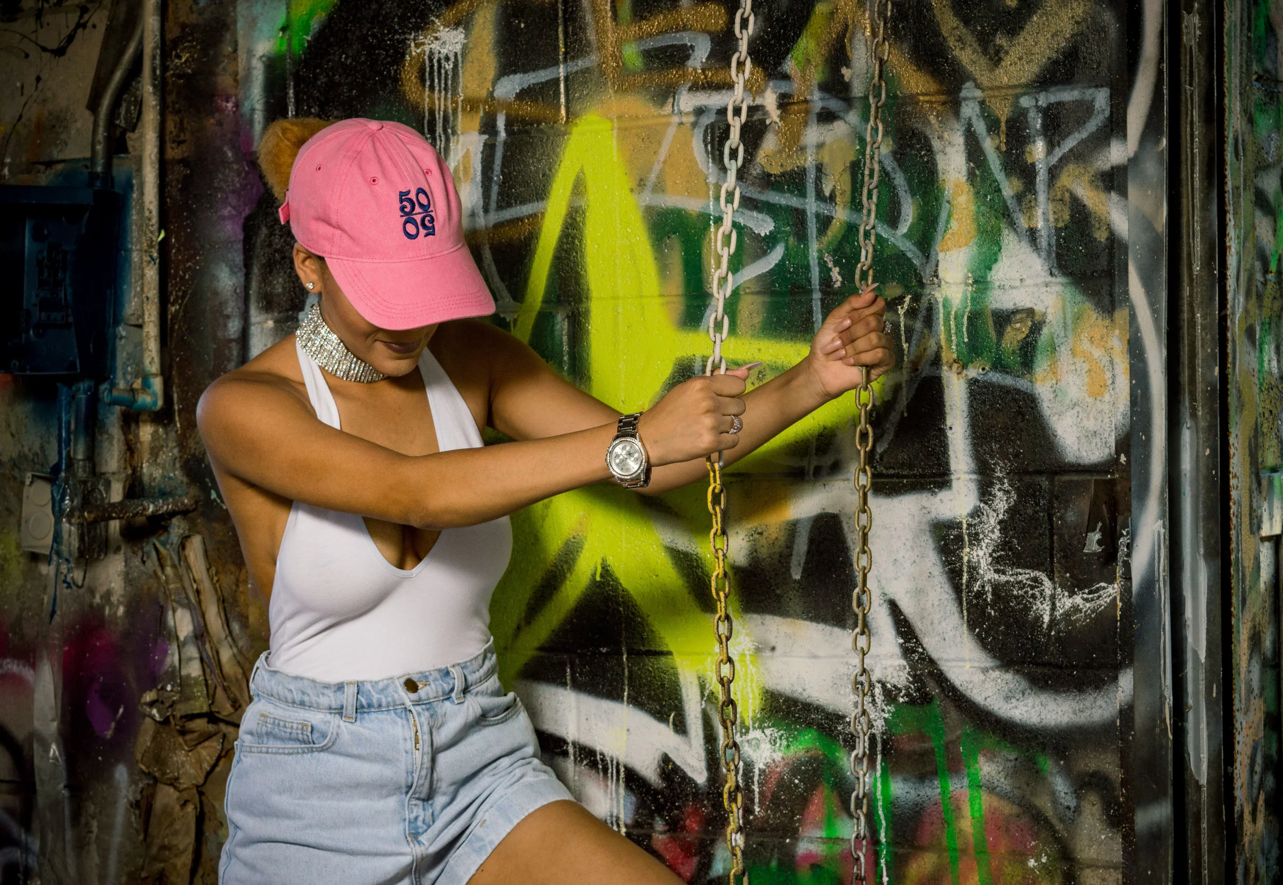  Female model wearing a pink trucker hat with 50/50 logo, standing in front of a graffiti wall, holding onto chains while wearing a white top and high-waisted shorts 