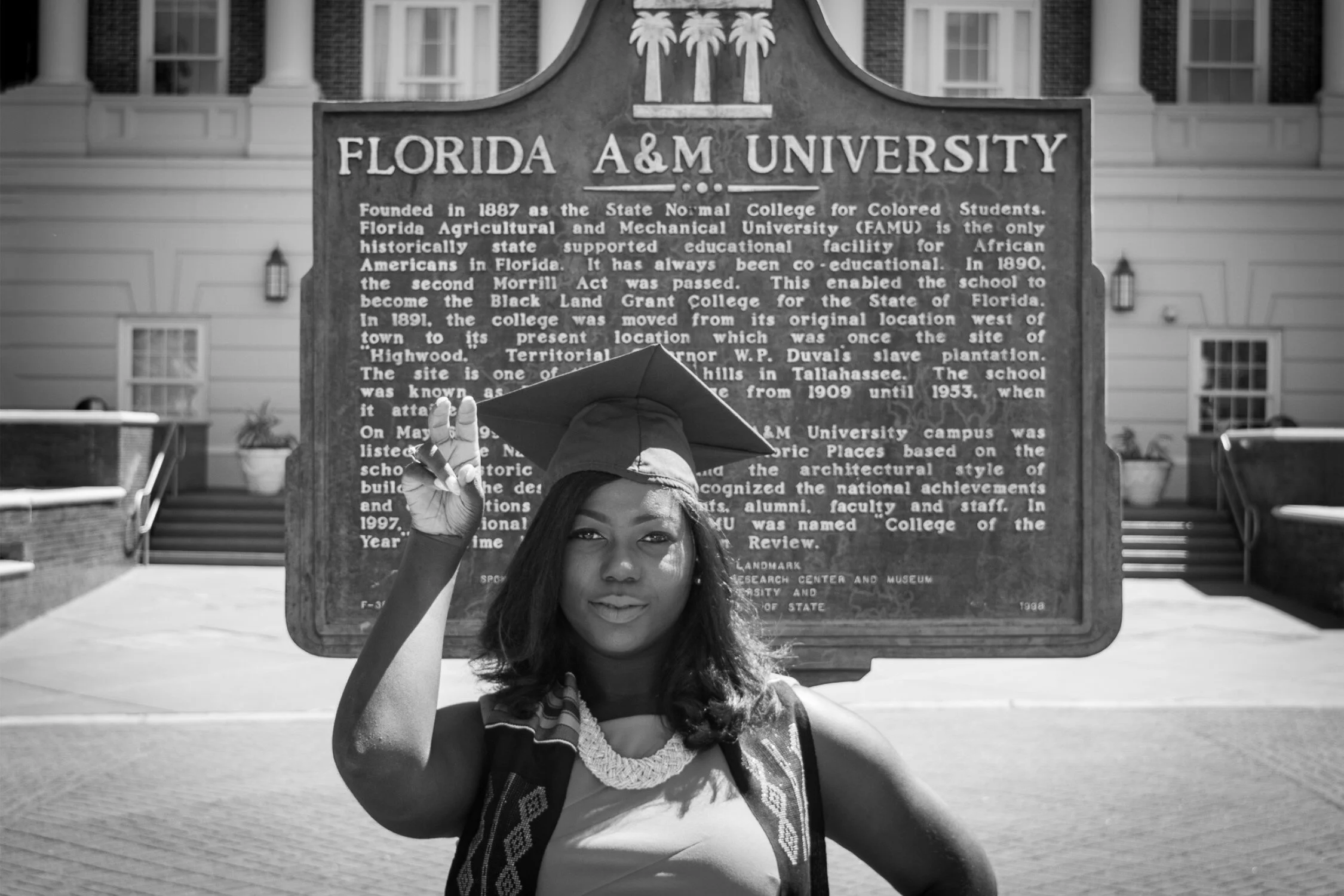  Black and white photo of FAMU graduate Shaperlya in front of Florida A&amp;M University information plaque 