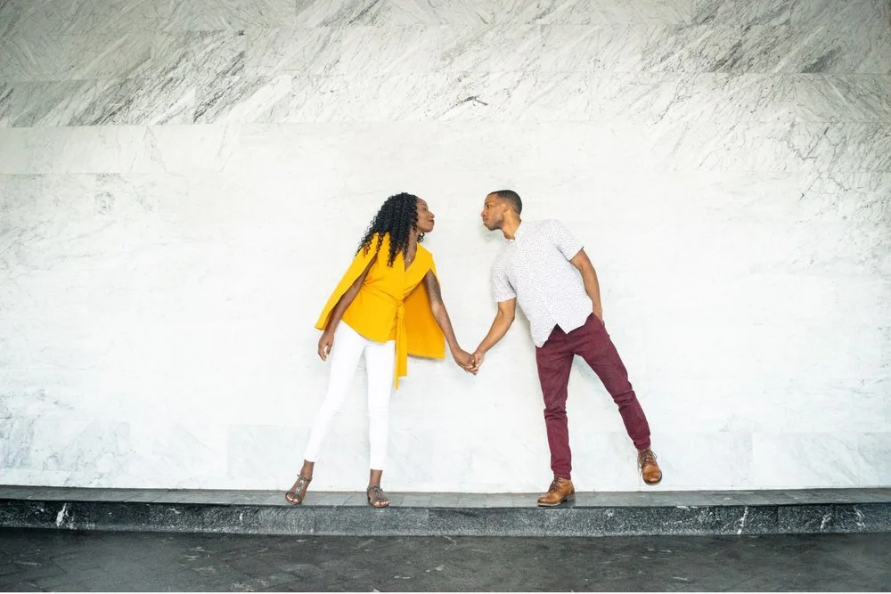  Unique engagement shoot photo of a couple leaning towards each other while attempting to balance on one foot. The couple leans their faces into one another as if about to kiss. 