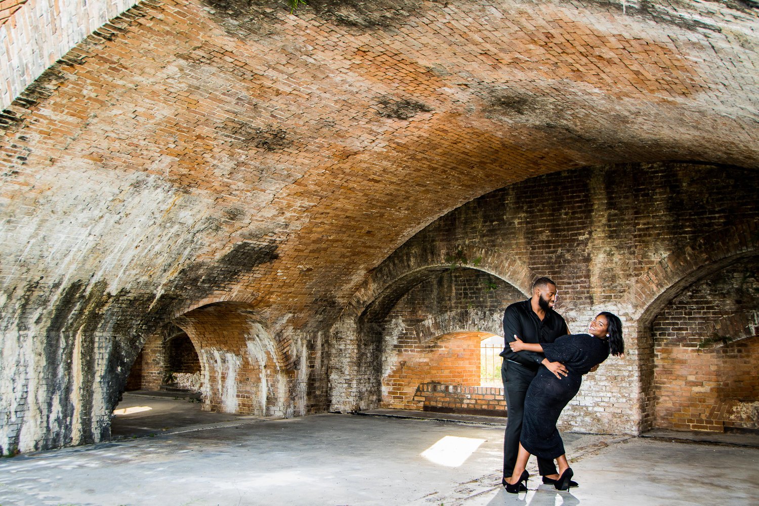  A couple dance in a large warehouse seeming space, wearing black formal wear for their engagement shoot. The man gazes at the woman as he dips her toward the floor, and the woman smiles at the camera. 