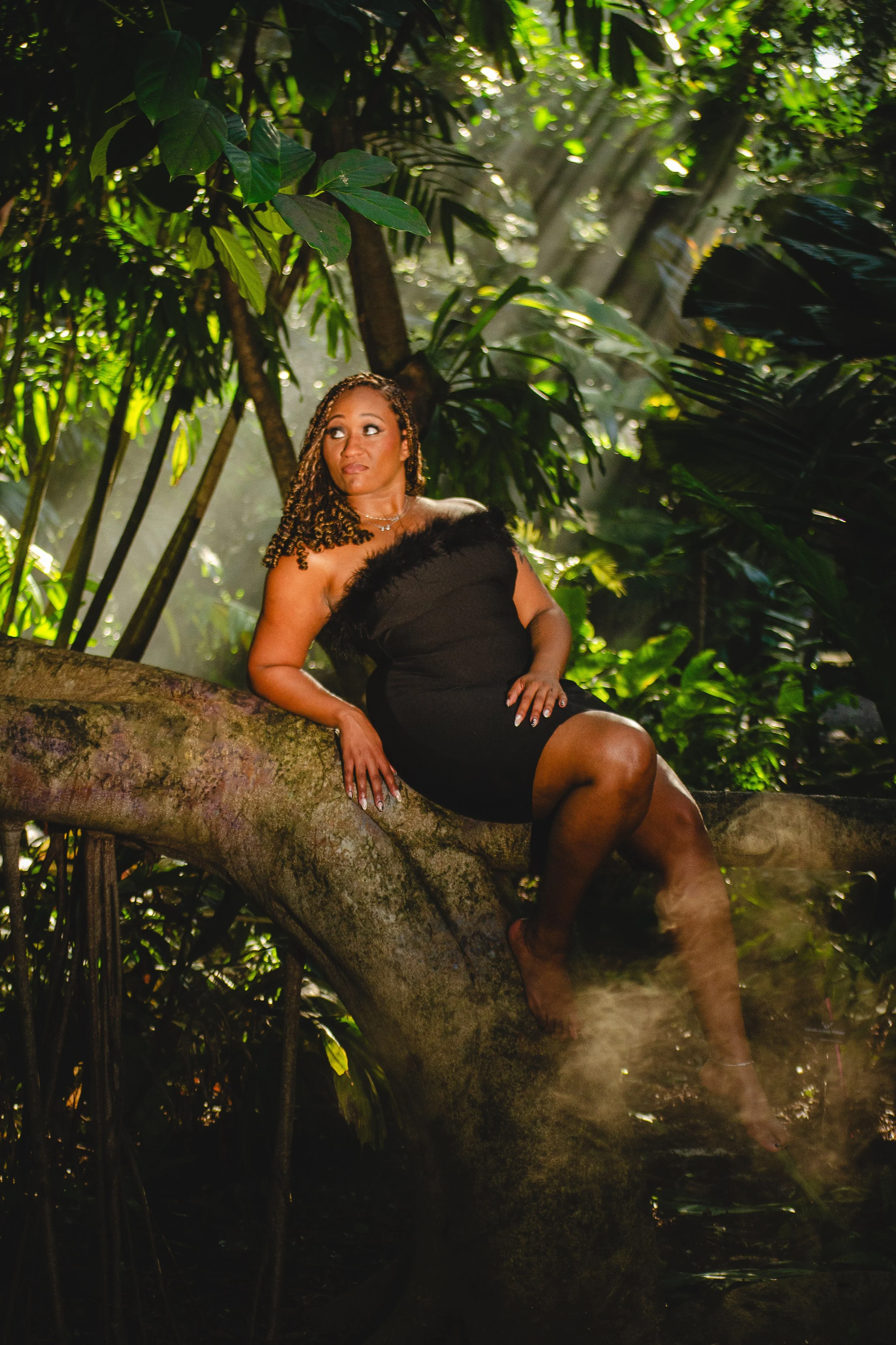  A woman in a strapless black dress with a feathered neckline sits gracefully on a large curved tree branch with one foot propped against the tree. She is surrounded by lush green foliage in a serene, tropical setting. Shafts of sunlight filter throu