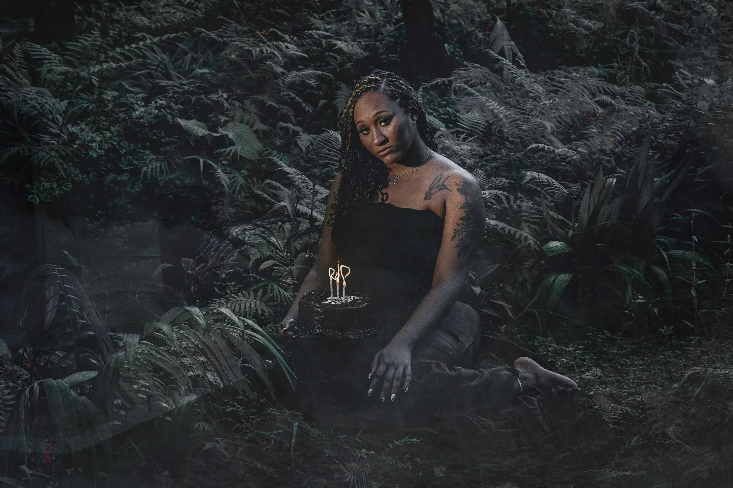  A woman in a black dress sits in dense moody foliage, holding a black cake with “RIP” candles glowing softly. The image has a dark, cinematic feel with desaturated tones, creating an eerie yet elegant atmosphere. 