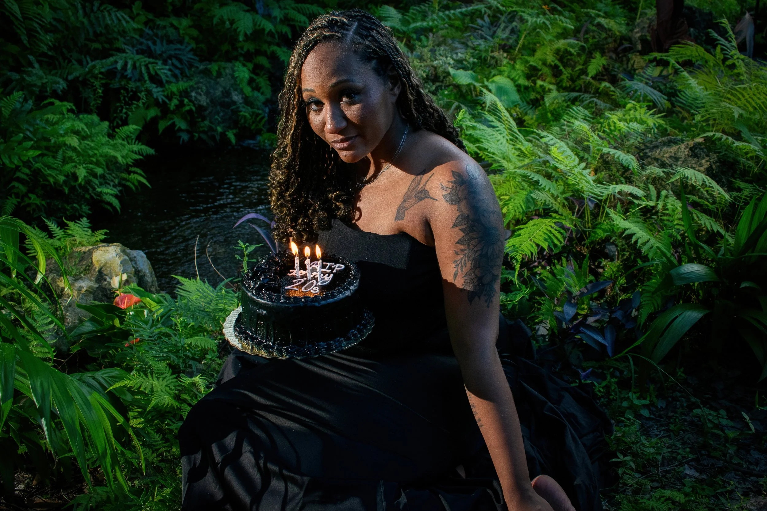 A woman in a black strapless dress sits in a lush, tropical setting, softly smiling and gazing at the camera, while holding a small black cake with white icing that reads “RIP to my 20s.” 