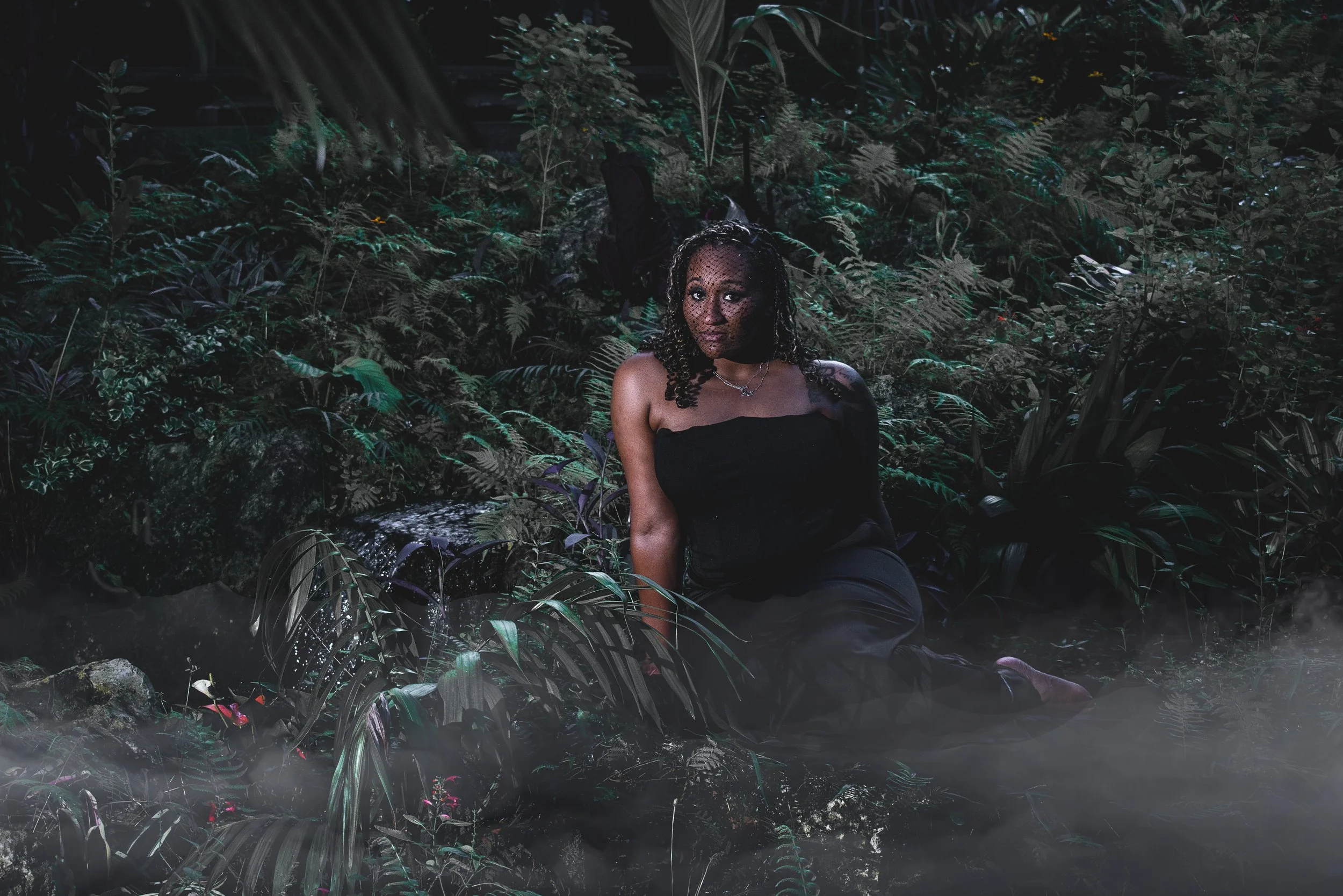  A woman in a black dress and black veil sits in dense moody foliage while leaning on her arm and gazing into the camera. The image has a dark, cinematic feel with desaturated tones, creating an eerie yet elegant atmosphere. 