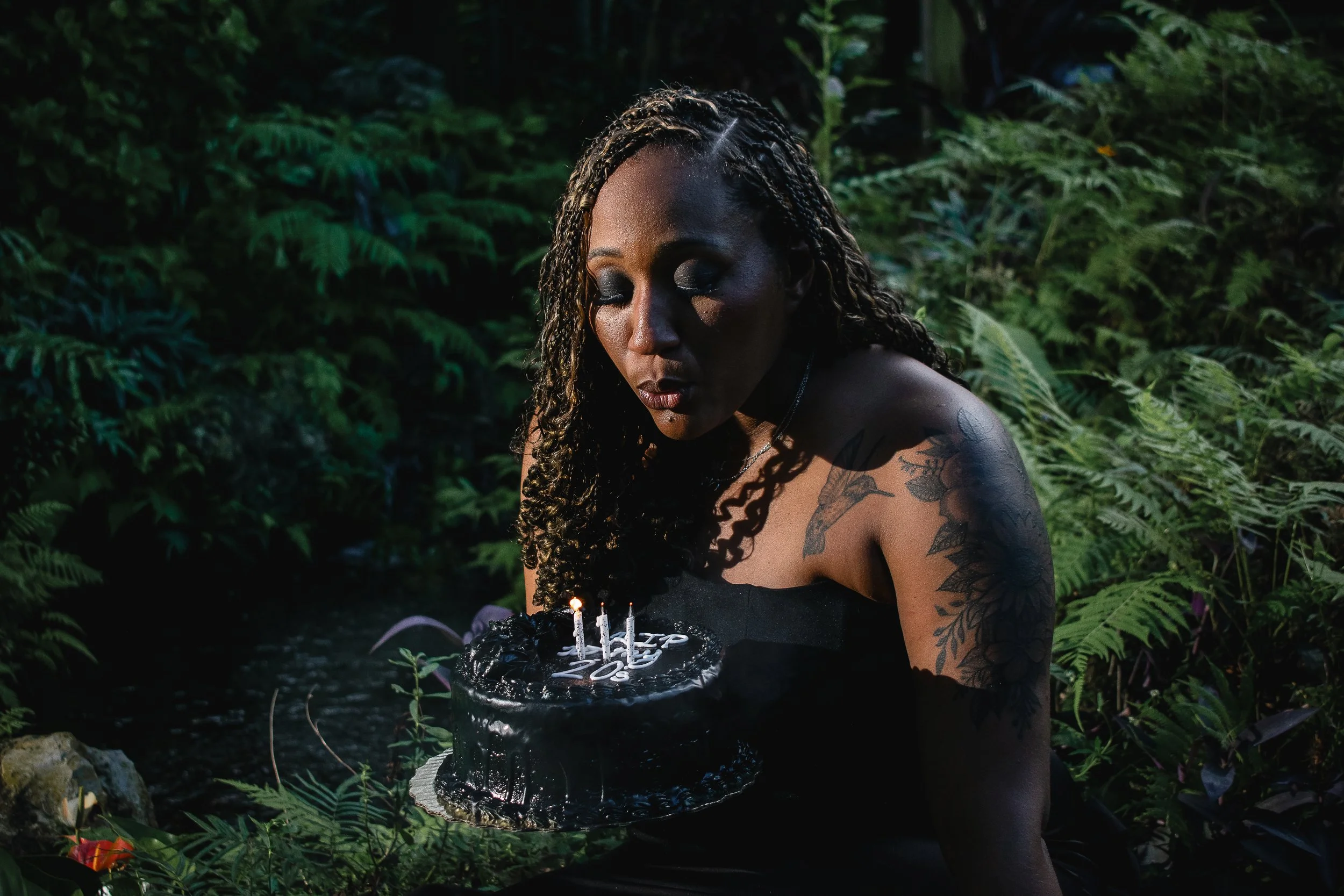  A woman in a black strapless dress sits in a lush, tropical setting, and prepares to blow out 3 candles, while holding a small black cake with white icing that reads “RIP to my 20s.” 