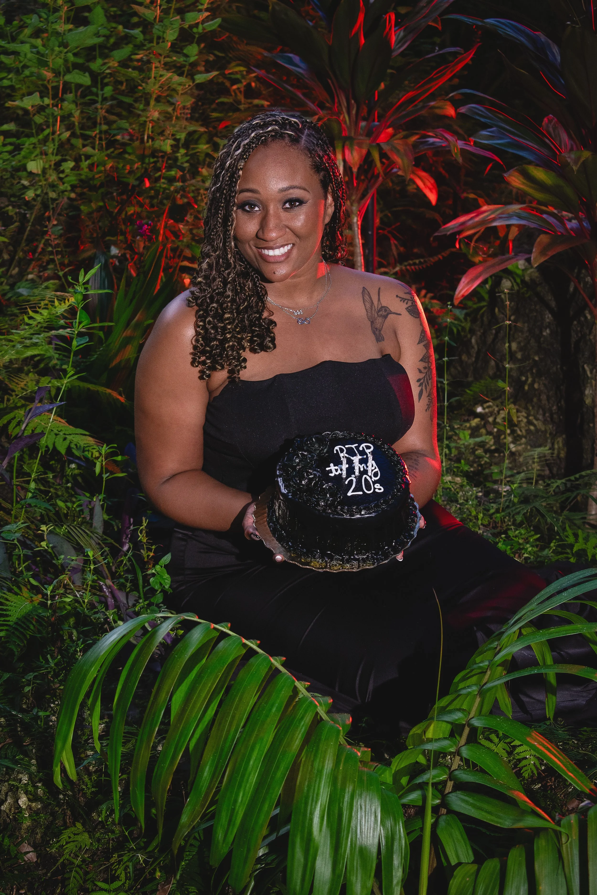  A woman in a black strapless dress sits in a lush, tropical setting, smiling while holding a small black cake with white icing that reads “RIP to my 20s.” The background is illuminated with warm red lighting, contrasting with the greenery surroundin
