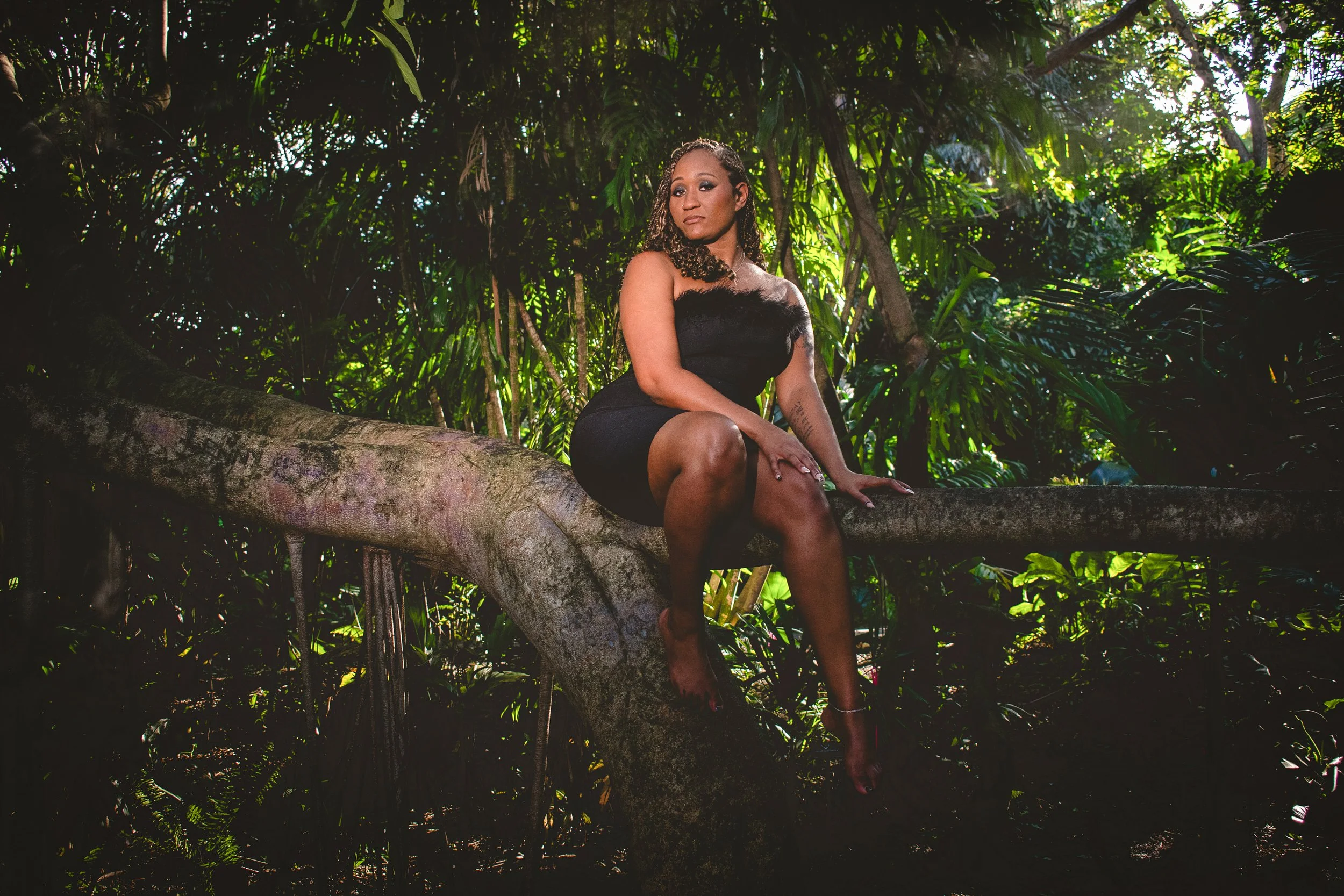 A lower-angled view of a woman in a strapless black dress with a feathered neckline who sits gracefully on a large curved tree branch with one foot propped against the tree. She is surrounded by lush green foliage in a serene, tropical setting. Shaf