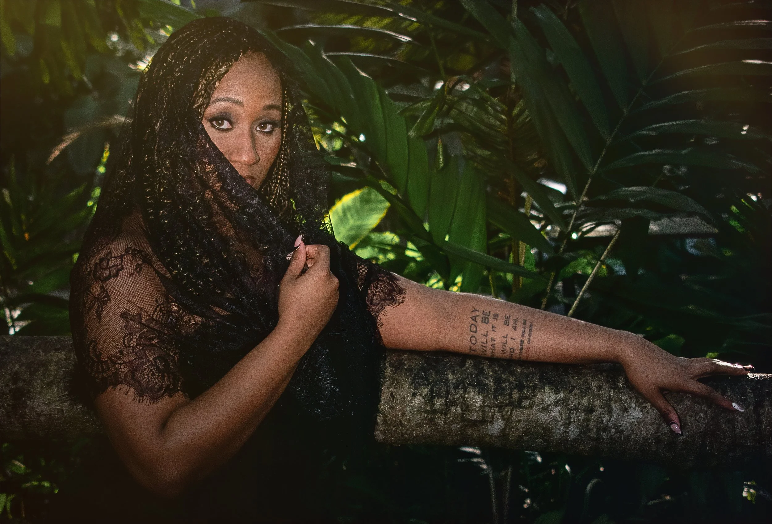  A close-up portrait of a woman draped in a black lace veil, partially covering her face as she gazes directly into the camera. Her arm rests on a large tree branch, framed by vibrant green tropical leaves in the background, with soft sunlight enhanc