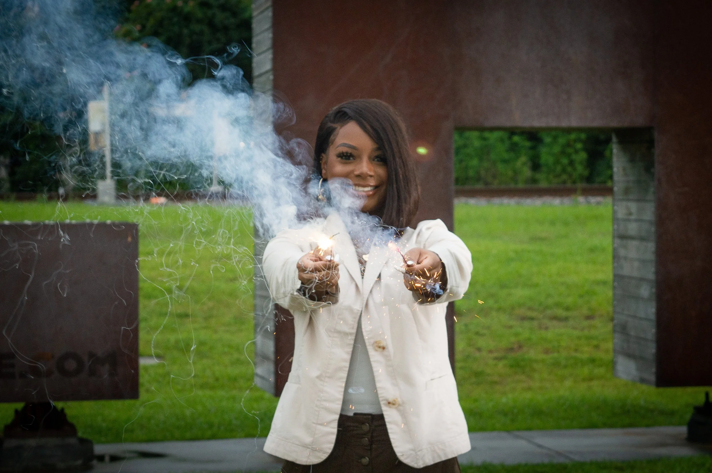 A young Black woman wearing a cream-colored blazer holds two sparklers in front of her face and smiles.