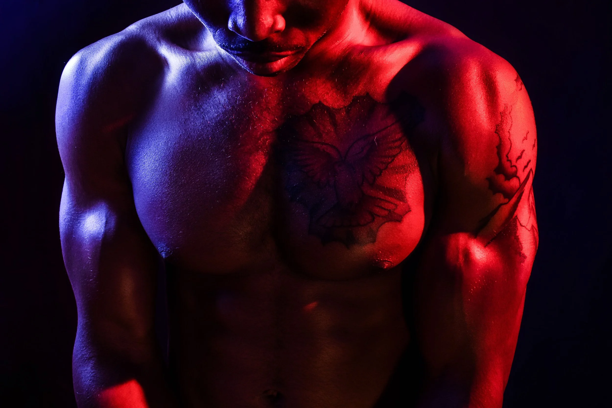 A male personal trainer poses in front of a black backdrop, wearing no shirt and bright yellow Gym Shark branded shorts. He flexes his biceps and pectoral muscles, which are illuminated and defined by the red and blue lights on both sides of him.