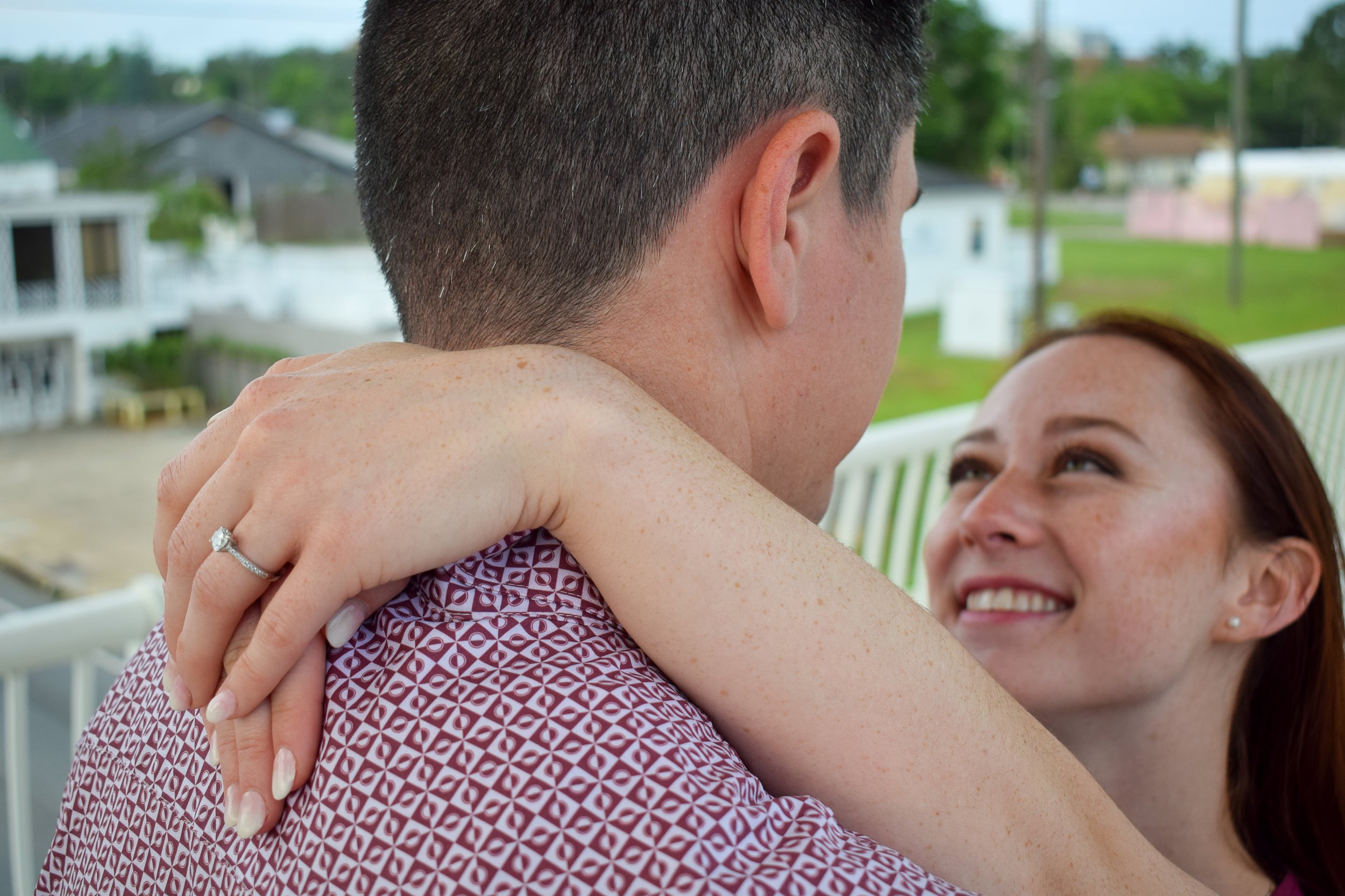 A woman smiles lovingly up into the face of her fiancé, with her arms draped around his shoulders. Visual focus is on the lovely engagement ring sparkling on her finger behind his head.