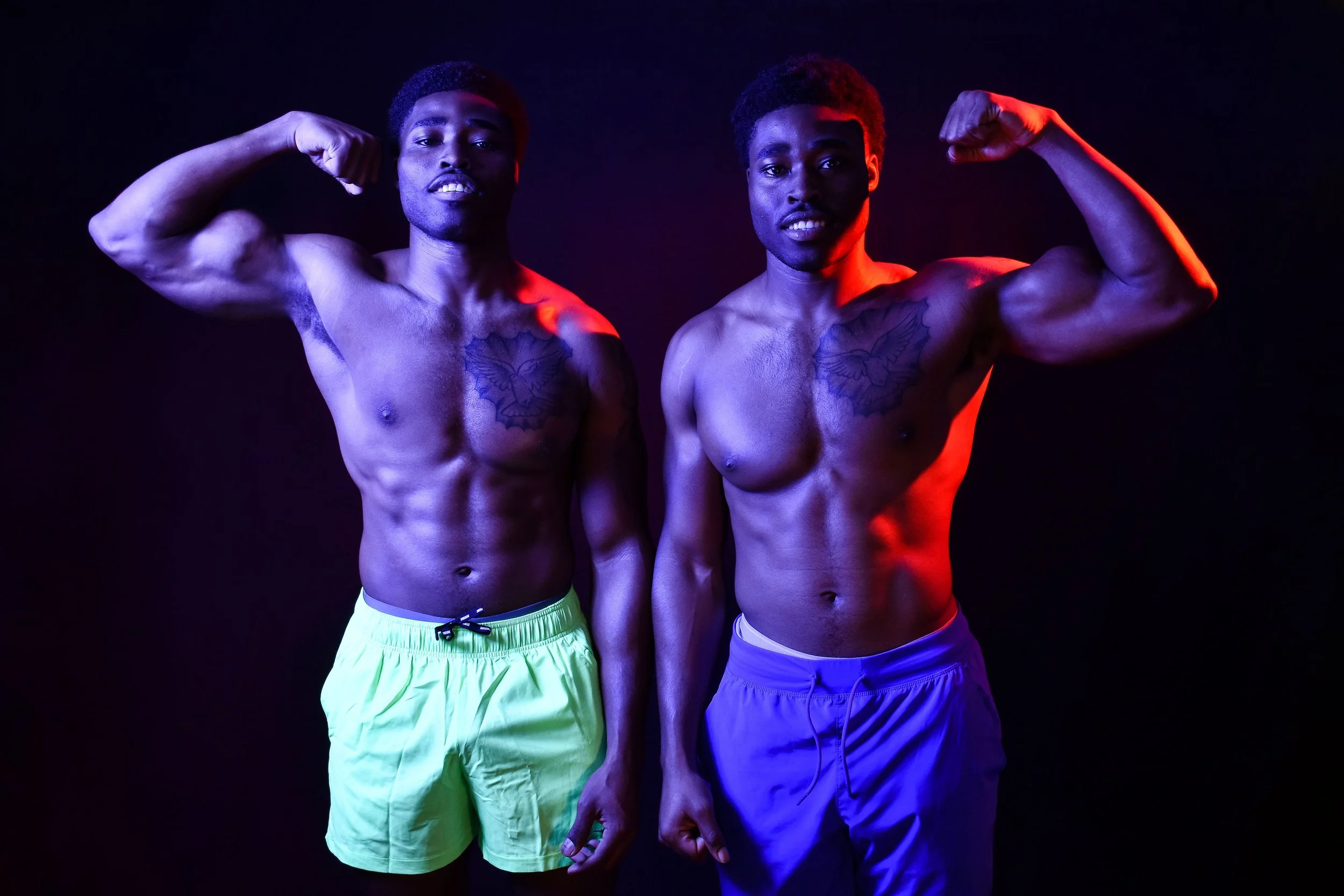 A male personal trainer poses with a client in front of a black backdrop, both wearing no shirt and Gym Shark branded shorts. They flex their muscles, which are illuminated and defined by the red and blue lights on either side.