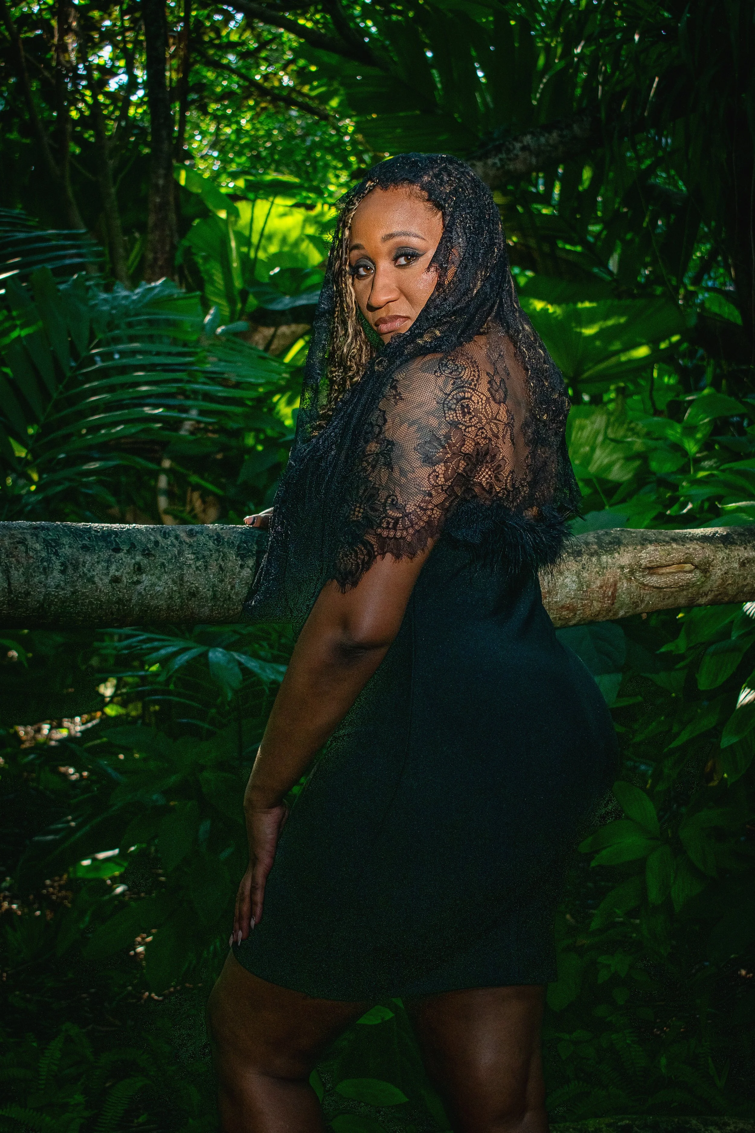 A woman wears a black dress and lace veil and looks over her shoulder at the camera with lush foliage of the Constitution Park Arboretum in the background.