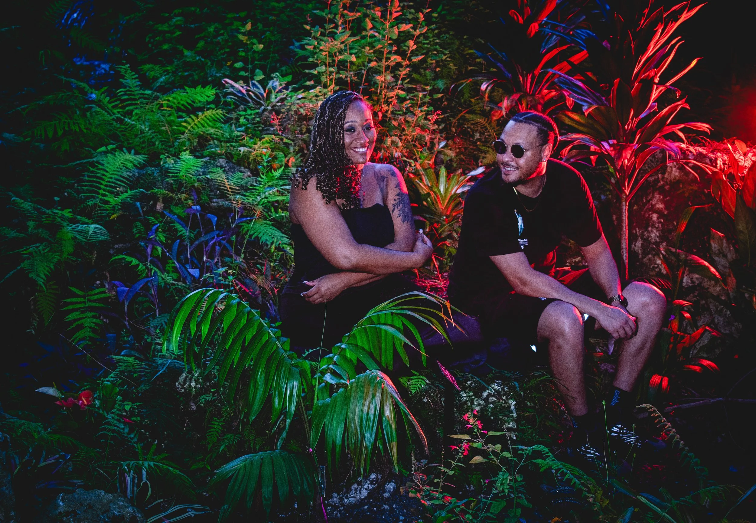 A couple laughs playfully while sitting by a small river surrounded by a colorful foliage. The couple are backlight by strong red lighting.