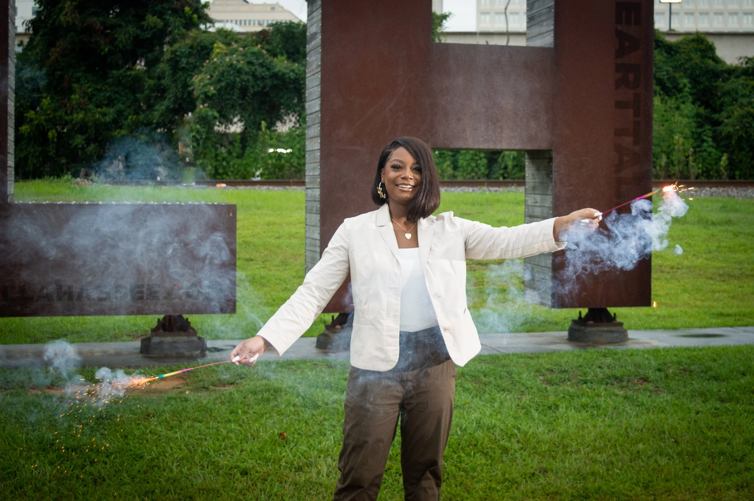 A young Black woman wearing a cream-colored blazer and chocolate brown slacks, stands in front of large copper and wooden "TLH"  letters, holding 2 sparklers and smiling.