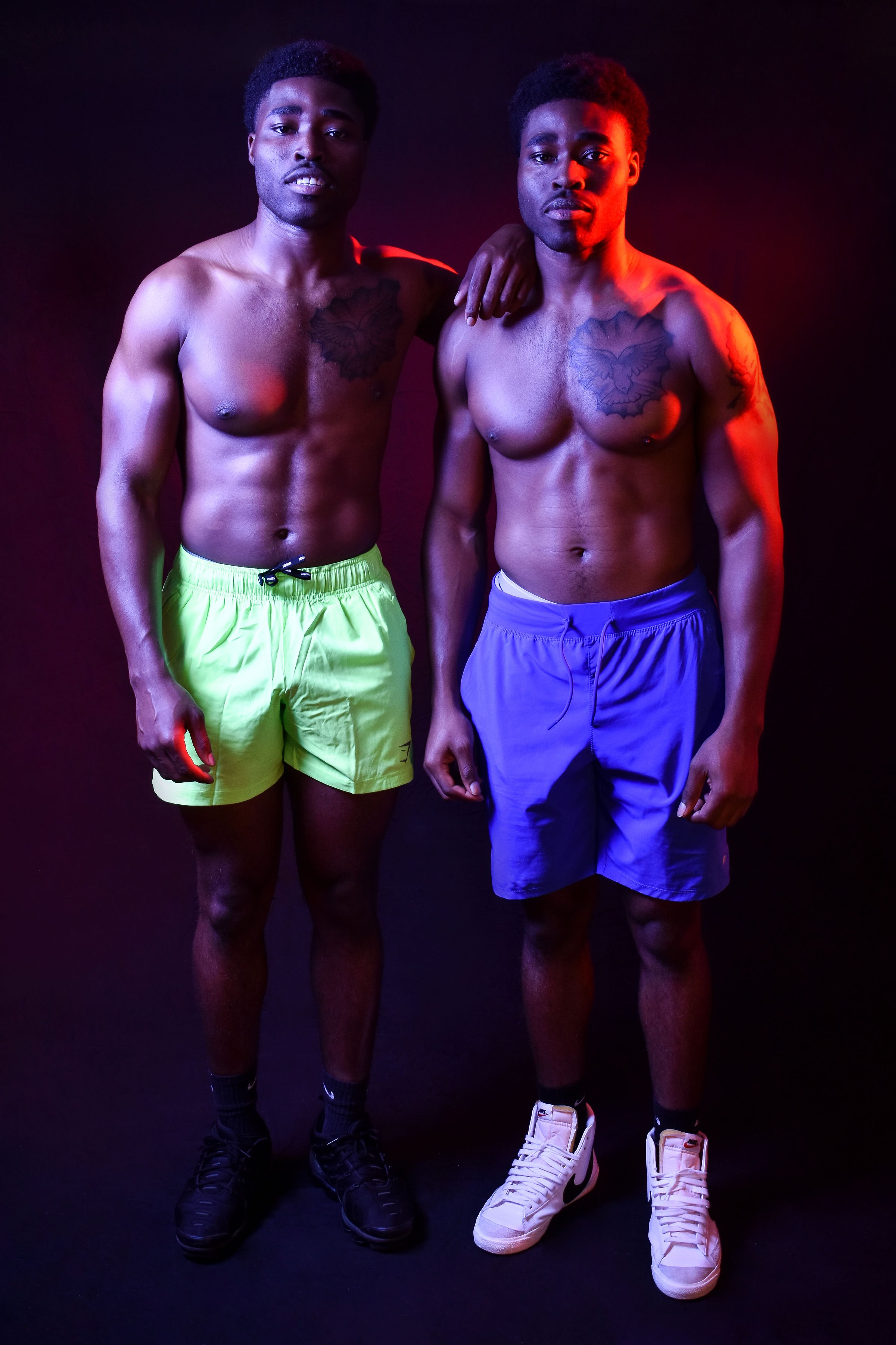 A male personal trainer poses with a client in front of a black backdrop, both wearing no shirt and Gym Shark branded shorts. They flex their muscles, which are illuminated and defined by the red and blue lights on either side.